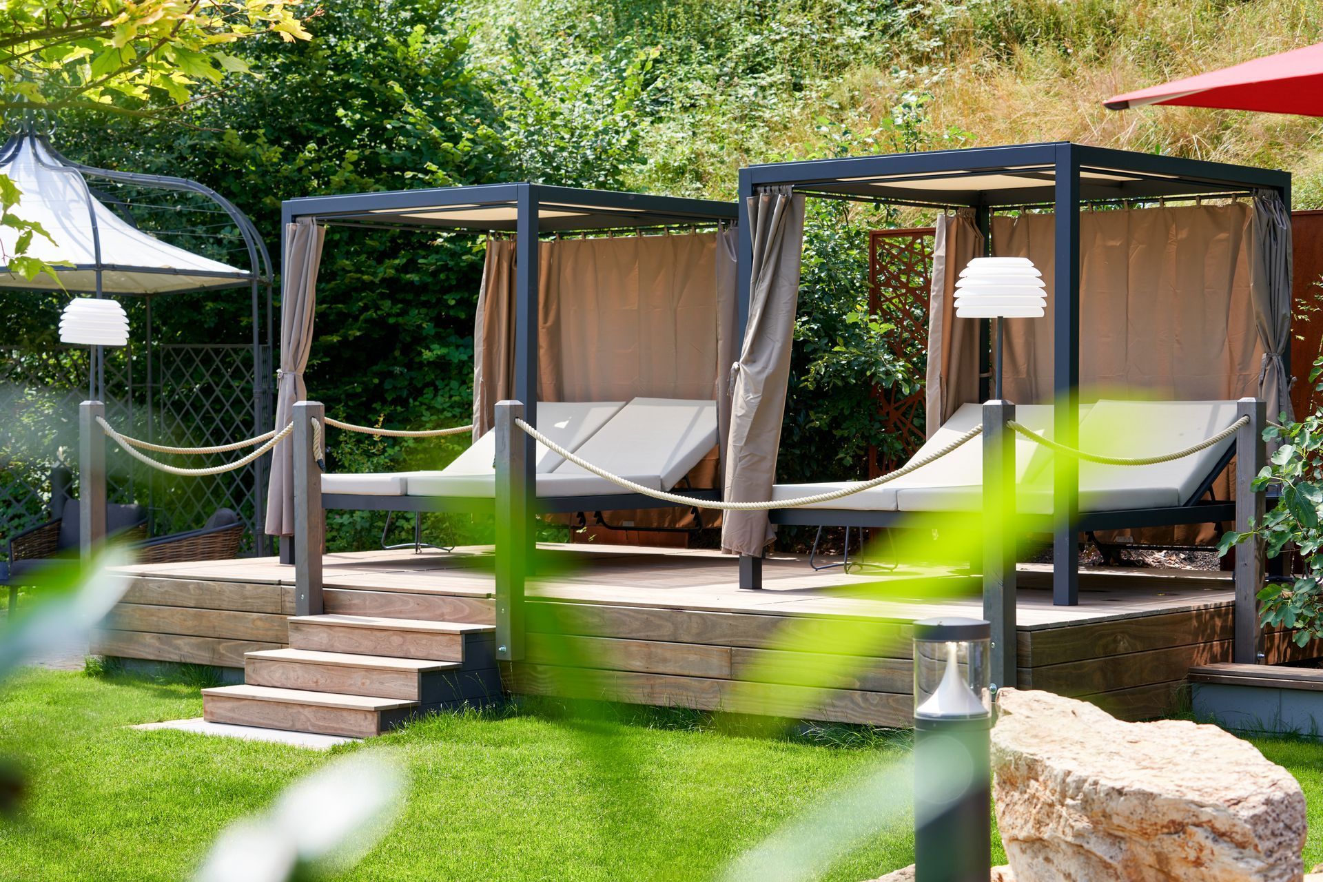Lounge im Freien mit dunklen Sonnensegeln, weißen Vorhängen und Stufen in einem üppigen Garten.