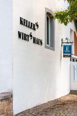 Weiße Gebäudewand mit dem Schild „KELLER'S WIRTS HAUS“ und einer blauen Hängetafel an einer Kopfsteinpflasterstraße.