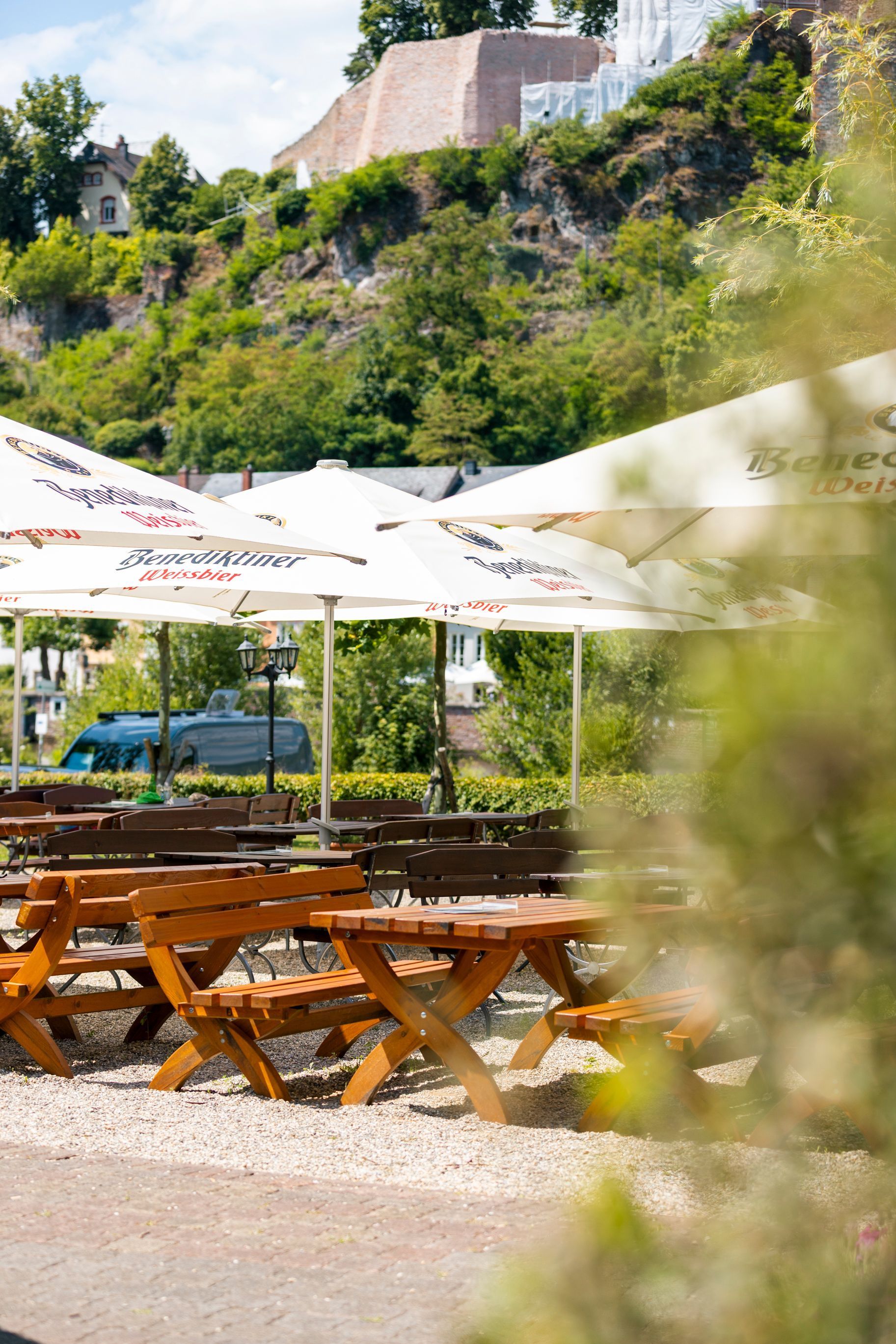 Café-Tische im Freien unter weißen Sonnenschirmen, im Hintergrund ein grüner Hügel und eine Steinmauer.