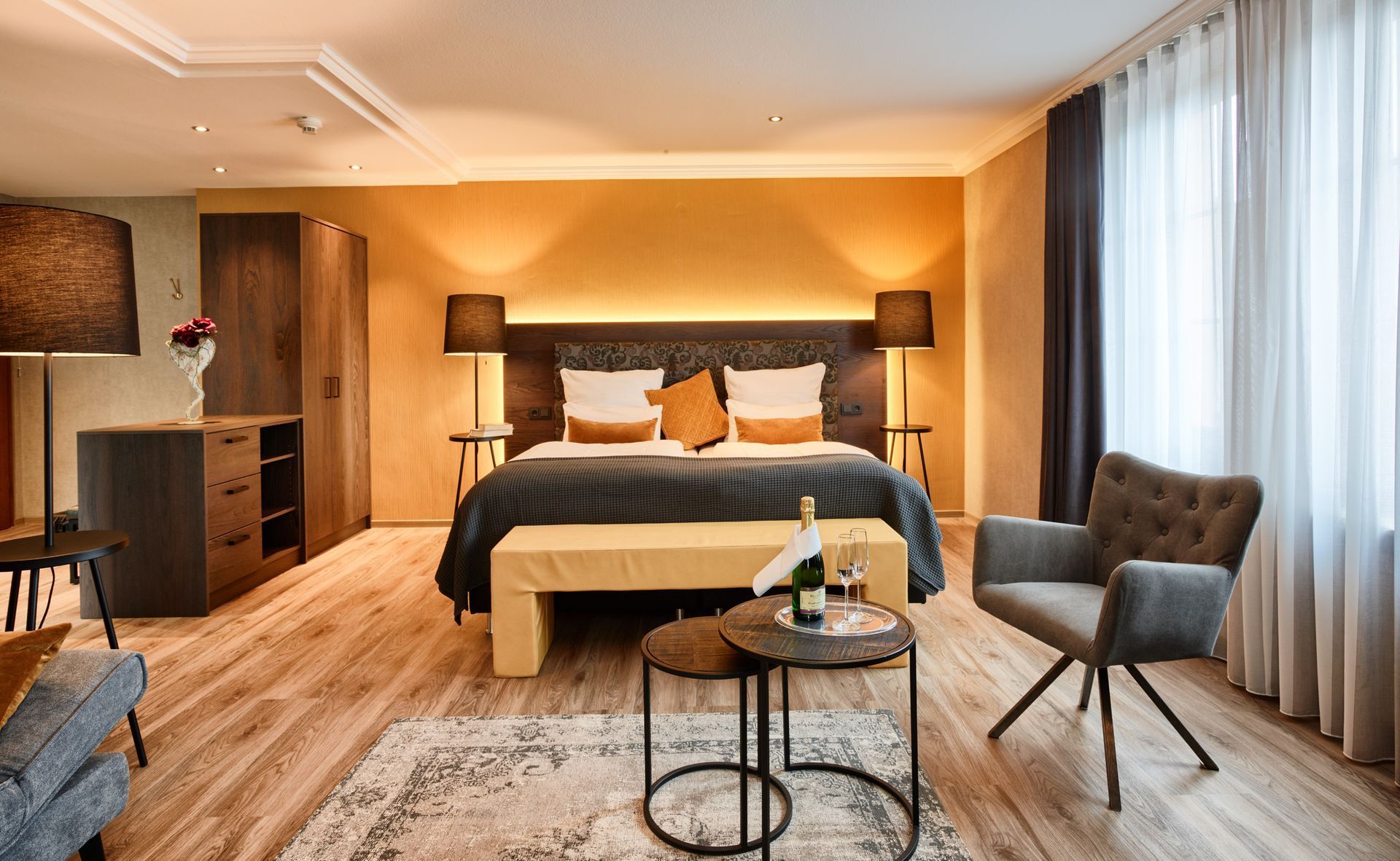 Modernes Schlafzimmer mit großem Bett, grauem Sessel, Couchtisch und bodenlangen Vorhängen