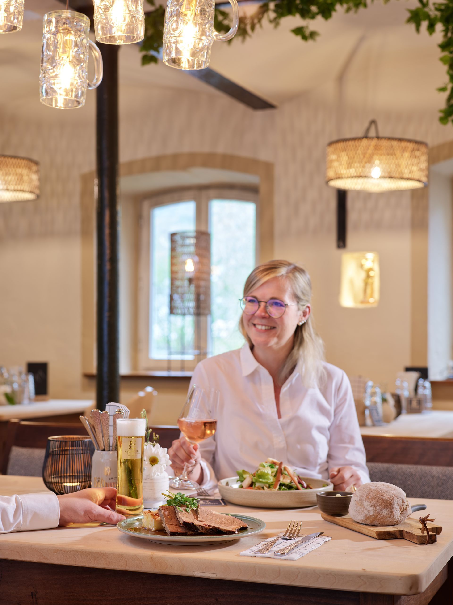 Eine lächelnde Person sitzt an einem Restauranttisch mit Speisen und Getränken unter warmem Licht.