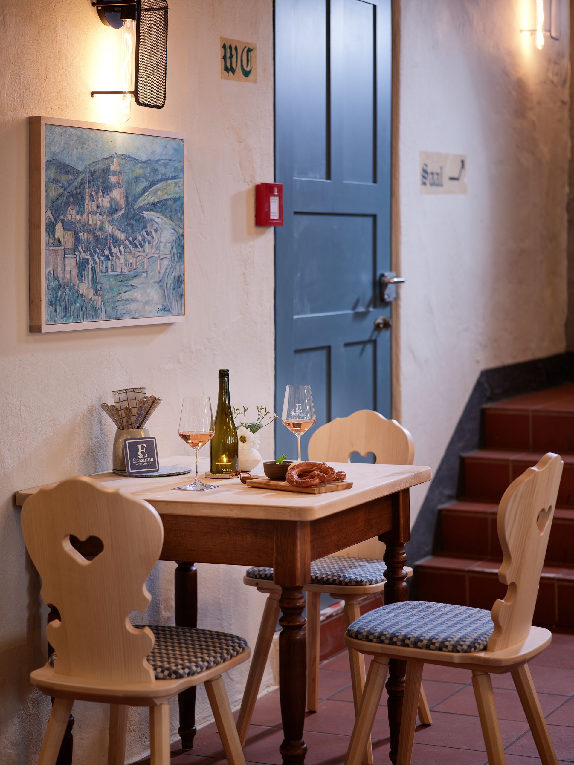 Kleiner Café-Tisch neben einer blauen Tür, mit zwei Holzstühlen, einer Weinflasche, Gläsern und einem gerahmten Wandbild.