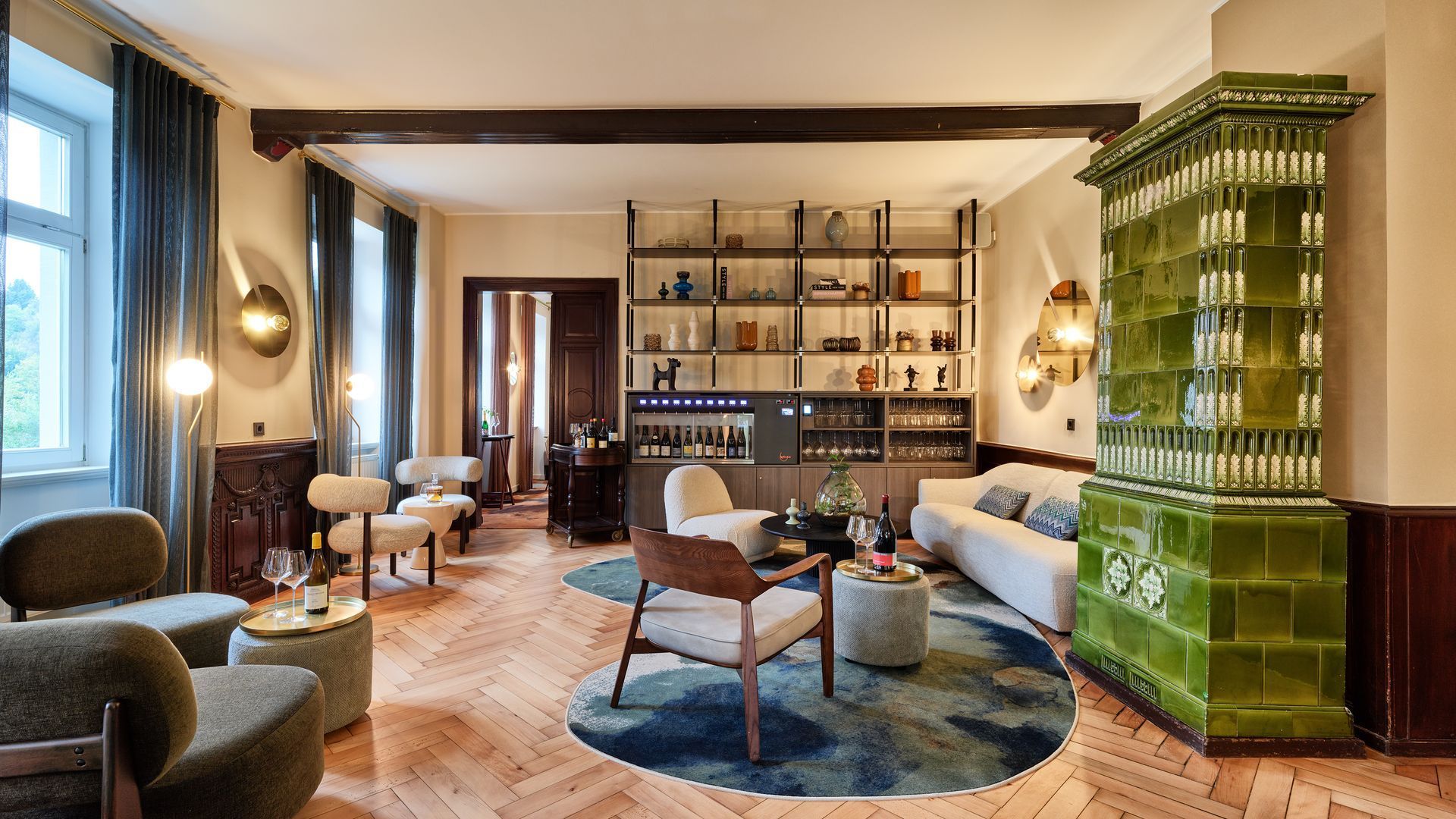 Stilvolles Wohnzimmer mit Sofas, einem blauen Teppich, Holzböden und einem grünen Kachelofen an großen Fenstern.