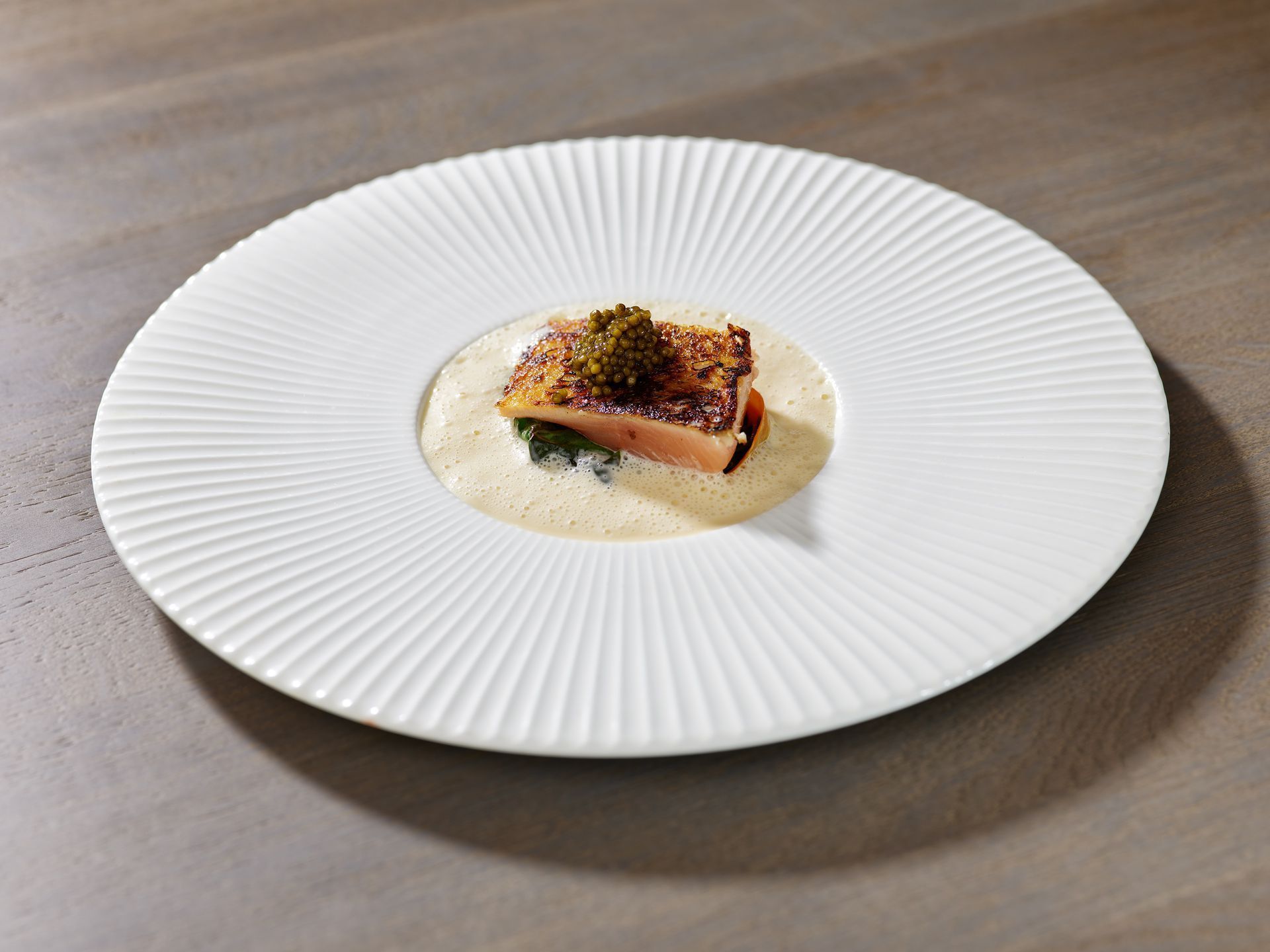 Ein Gourmet-Fischgericht mit cremiger Sauce, angerichtet auf einem weißen, geriffelten Teller, steht auf einem Tisch.