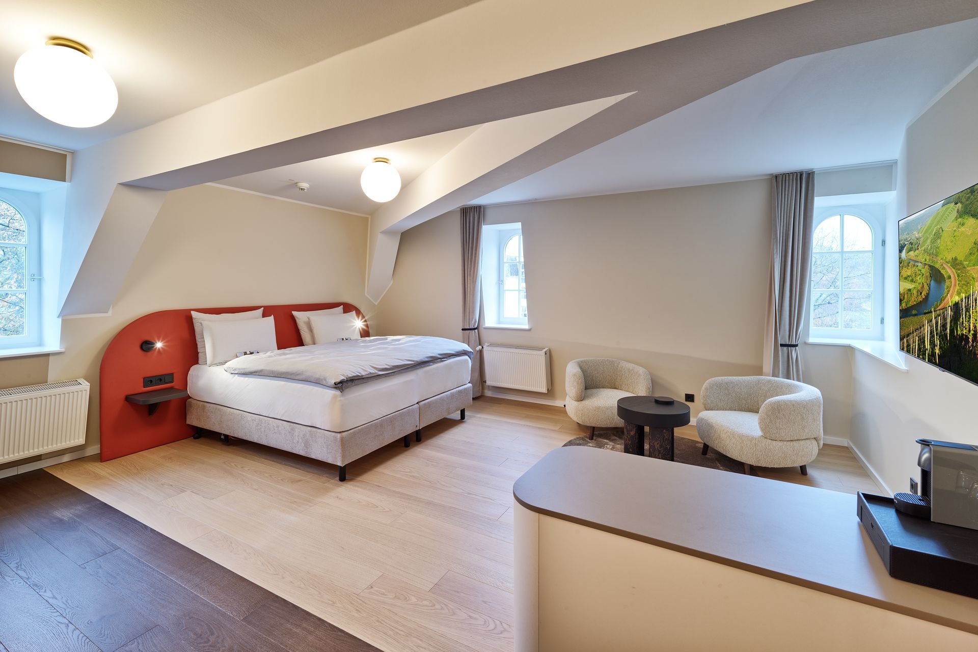 Helles Dachgeschoss-Schlafzimmer mit einem rot akzentuierten Bett, großen Fenstern und einem beigen Sessel an der Wand.