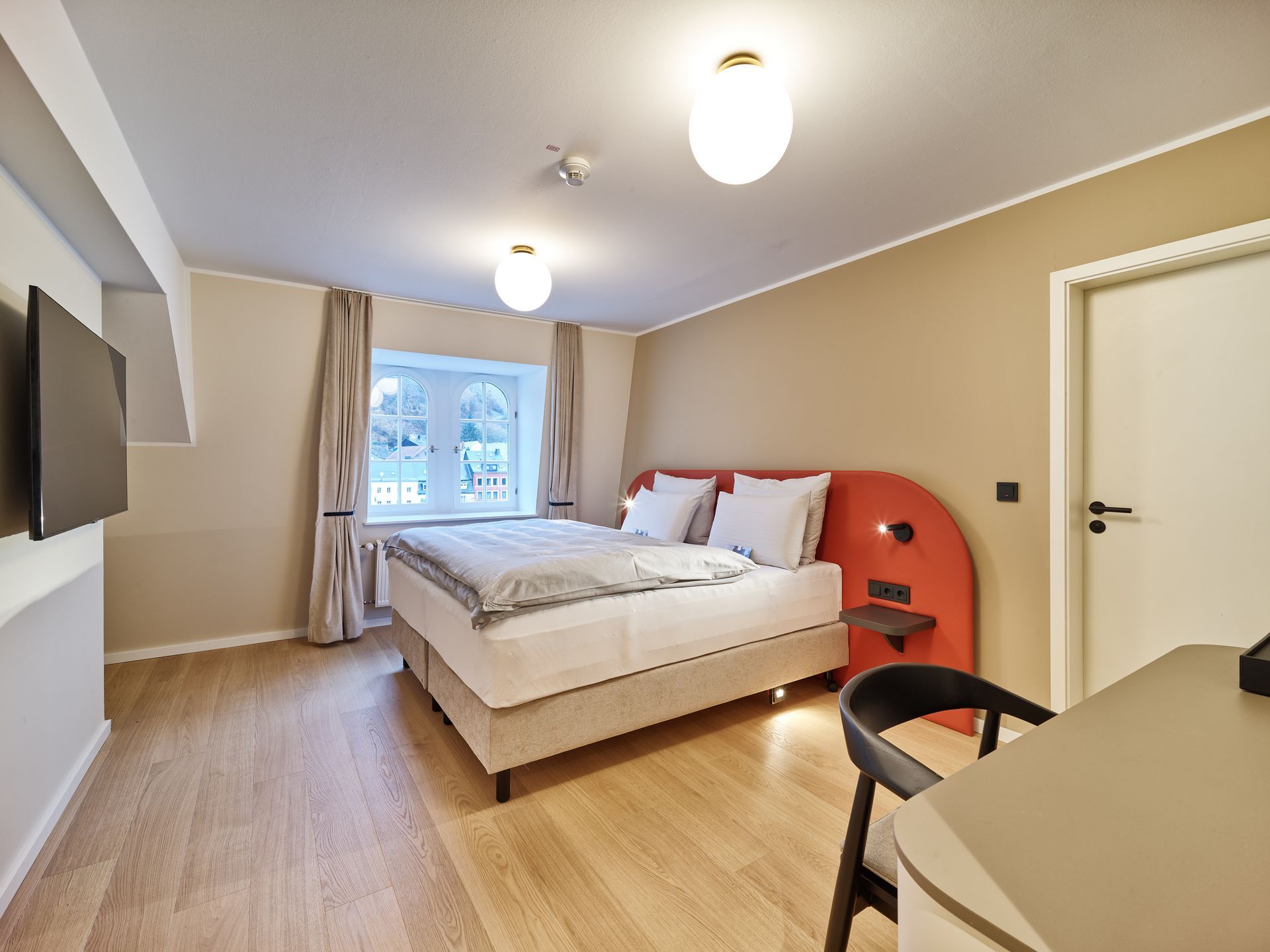 Gemütliches Schlafzimmer mit Bett, Fenster, beigen Wänden, Holzboden und einem kleinen Schreibtischstuhl auf der rechten Seite.