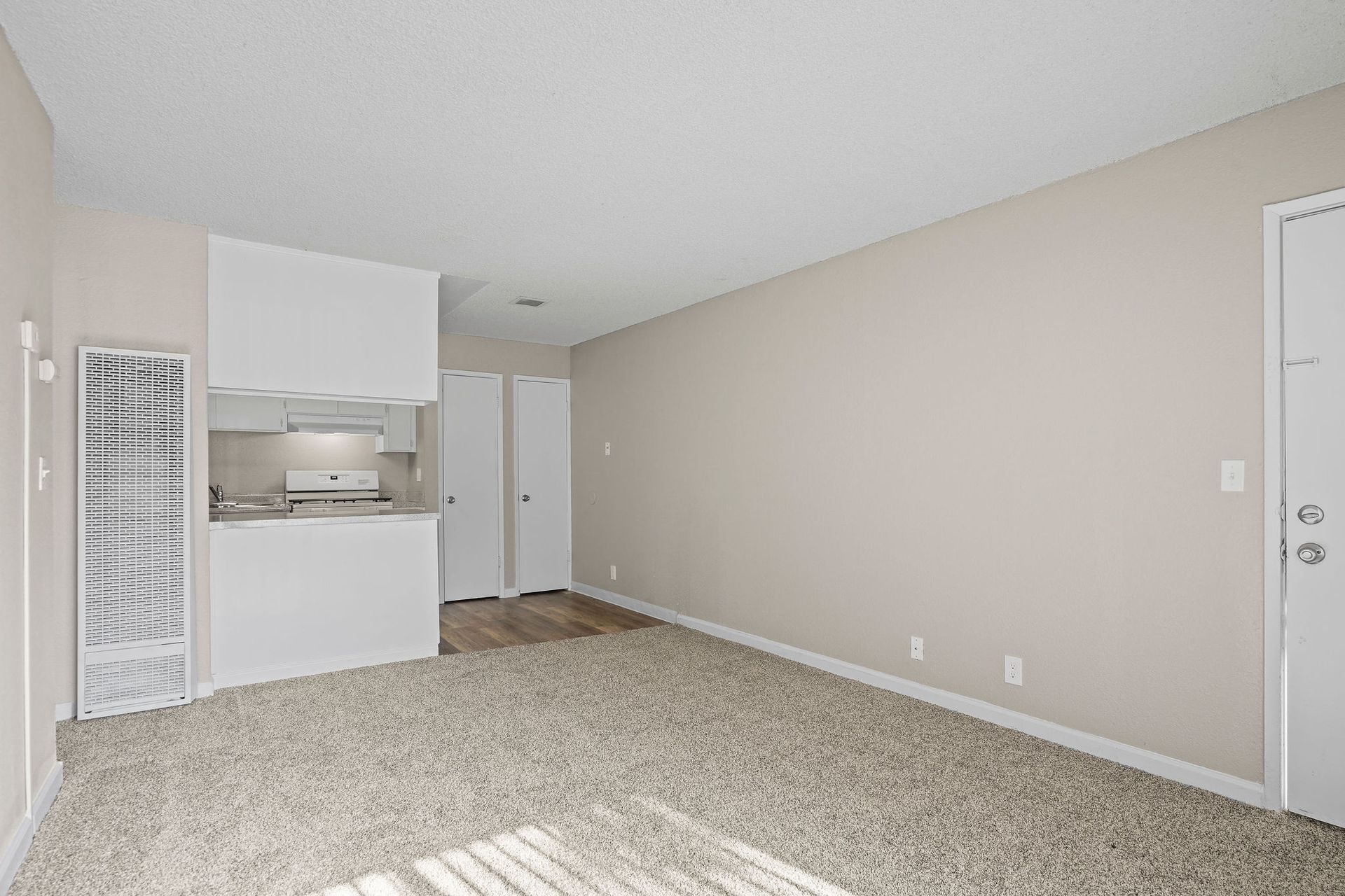 Empty beige carpeted living room with white kitchen area and sliding glass door blinds