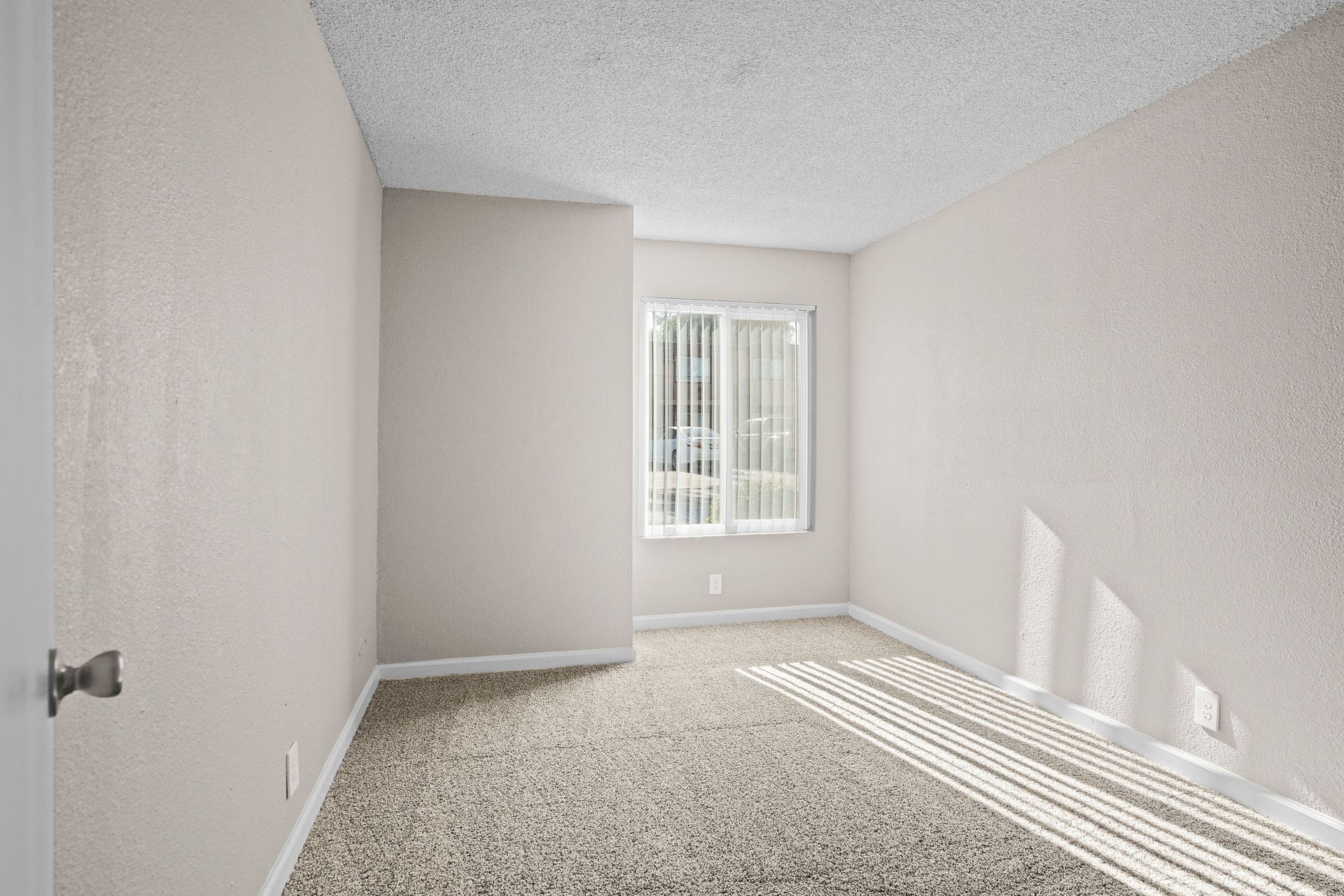 Empty beige bedroom with carpet, window blinds, and sunlight streaming across the floor