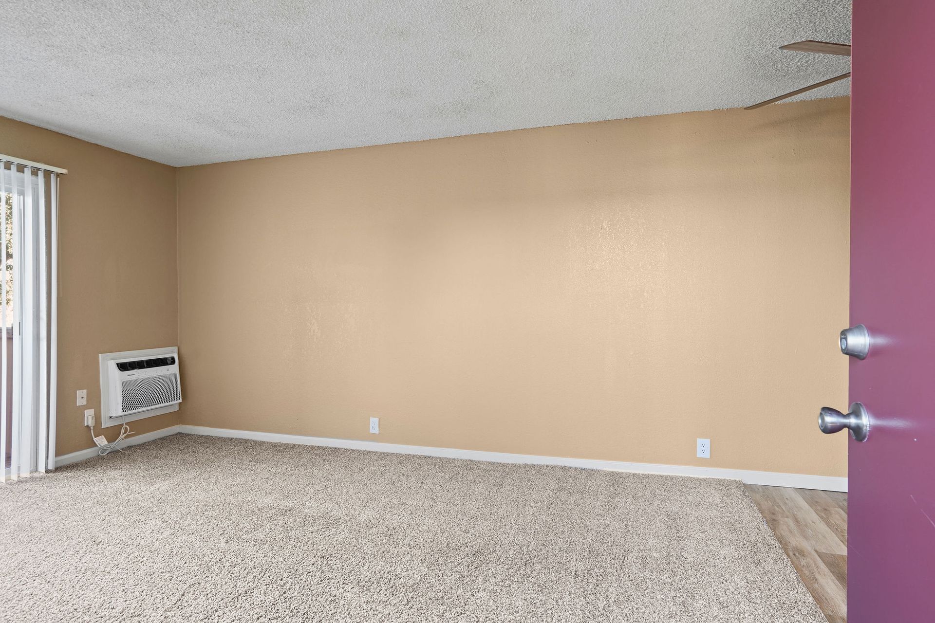 Empty beige-carpeted room with tan walls, a window, and a wall-mounted heater.