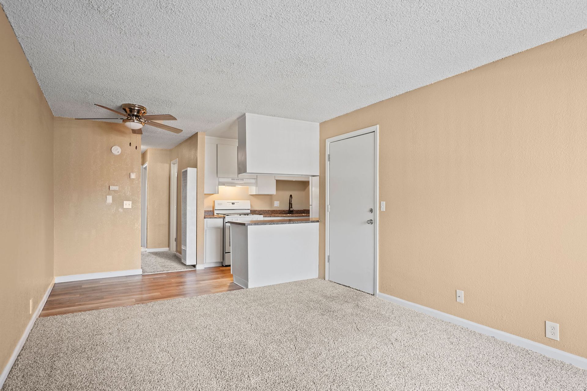 Empty beige living room with carpet, ceiling fan, and small kitchen area with white cabinets.