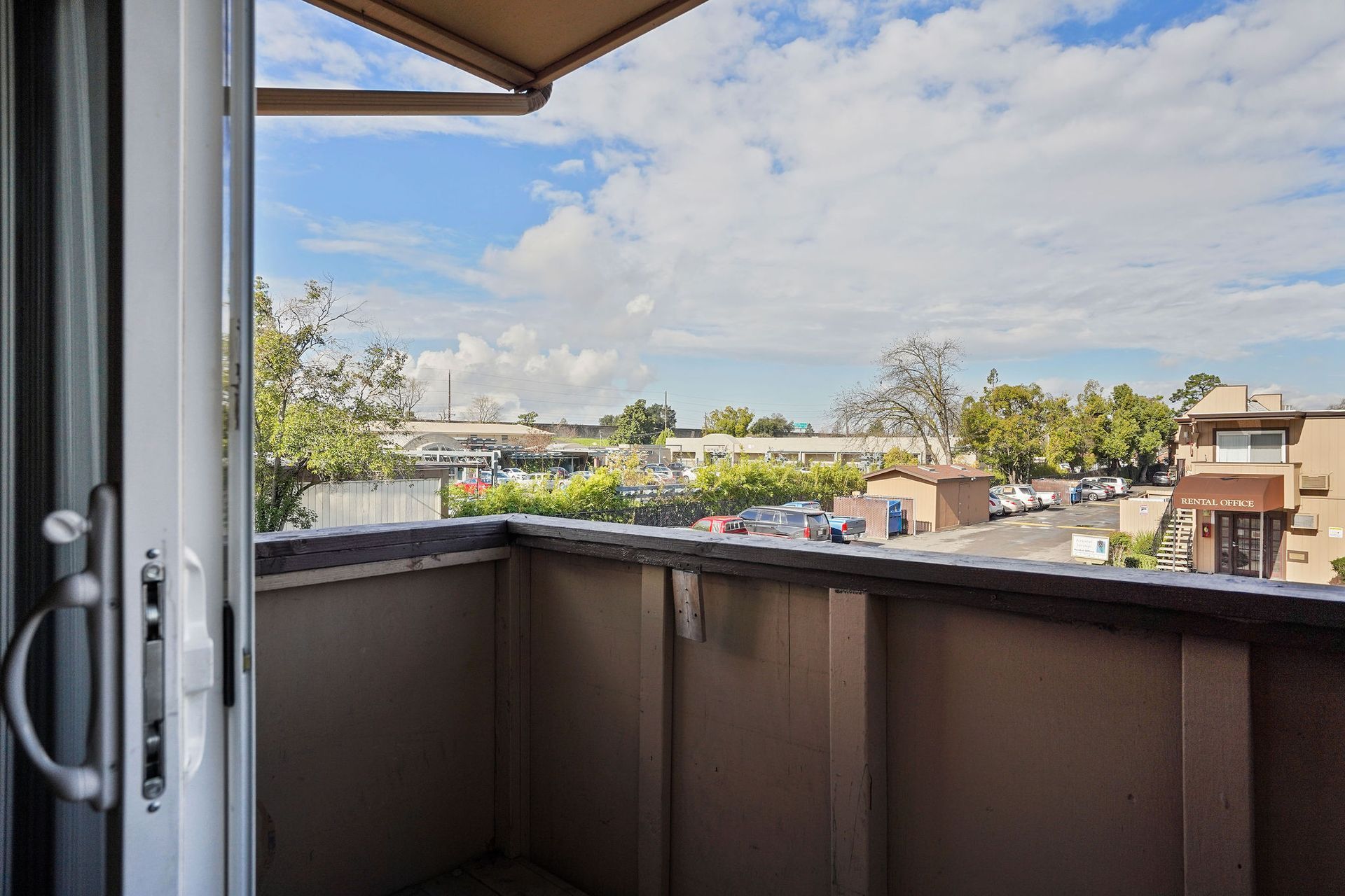 View from a balcony with dark railing, overlooking rooftops and trees under a partly cloudy sky.