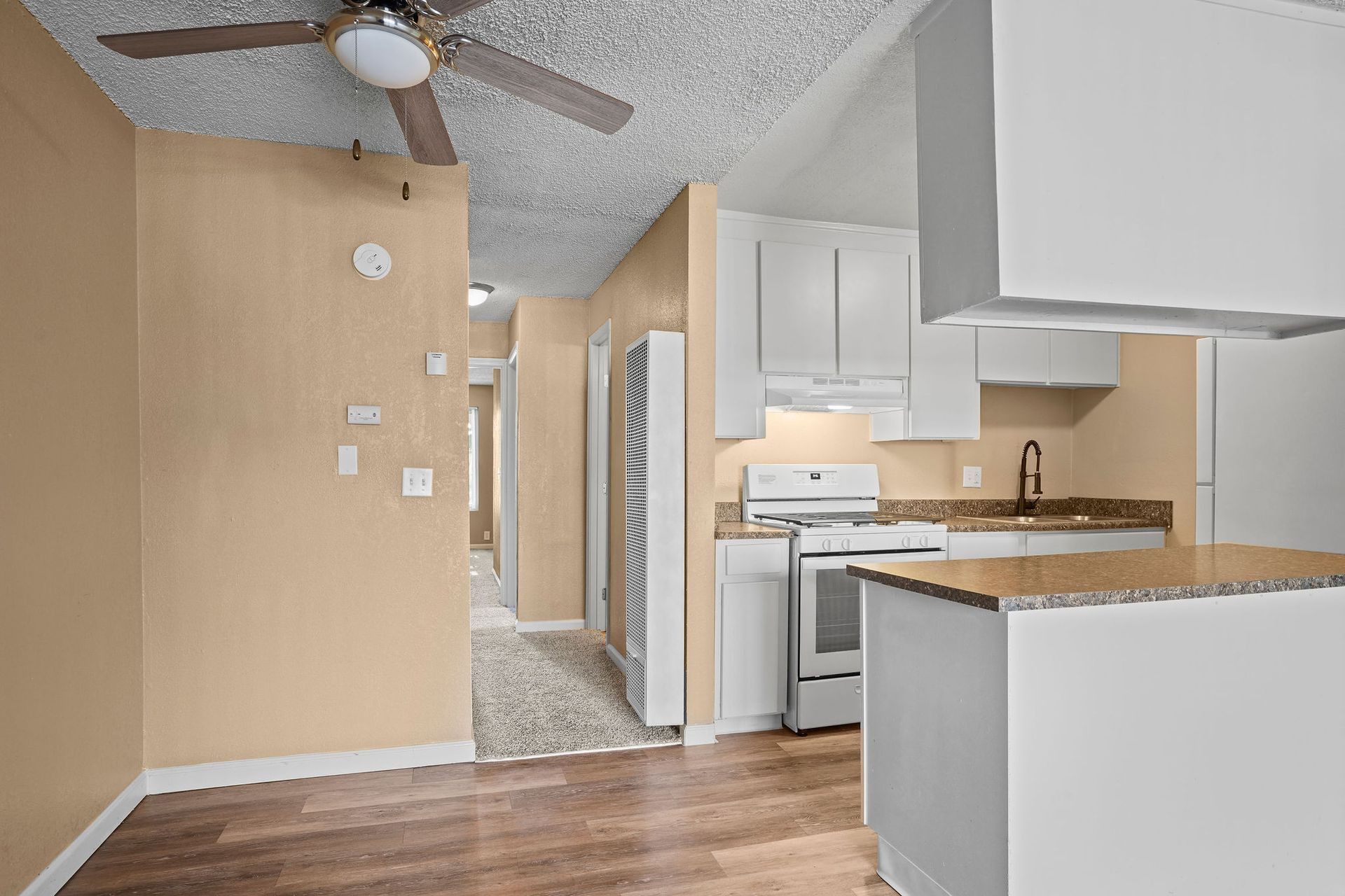 Empty kitchen with white cabinets, wood counters, ceiling fan, and hallway visible beyond.