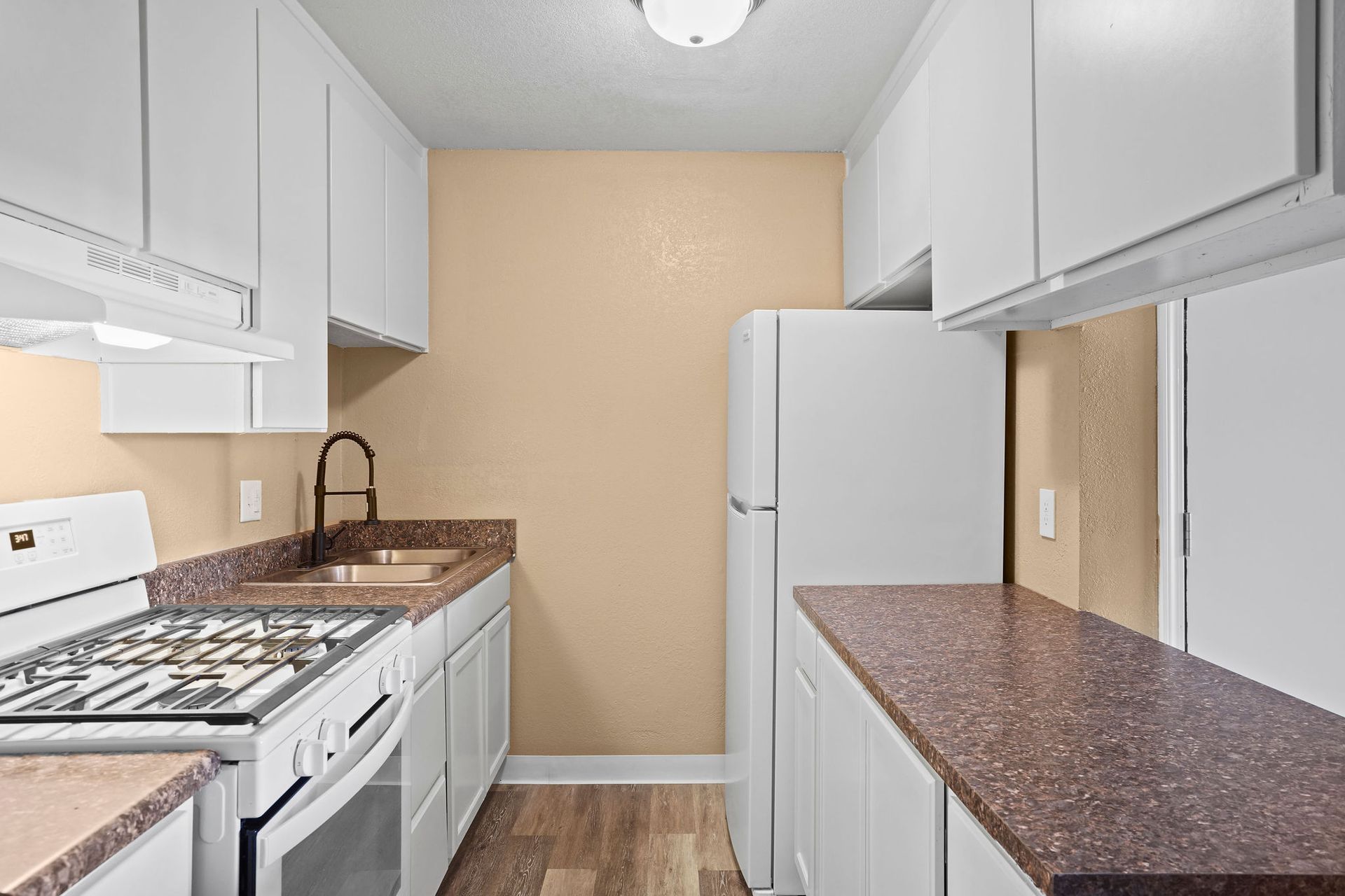 Narrow white kitchen with brown countertops, white cabinets, and a refrigerator against a beige wall.