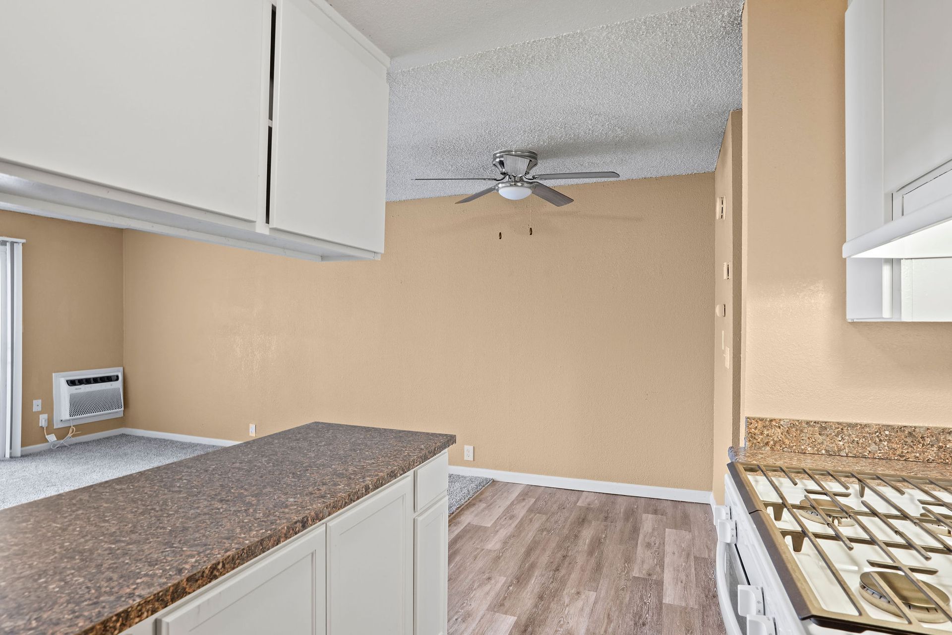 Empty kitchen with white cabinets, speckled countertops, gas stove, and beige walls leading to a ceiling fan area