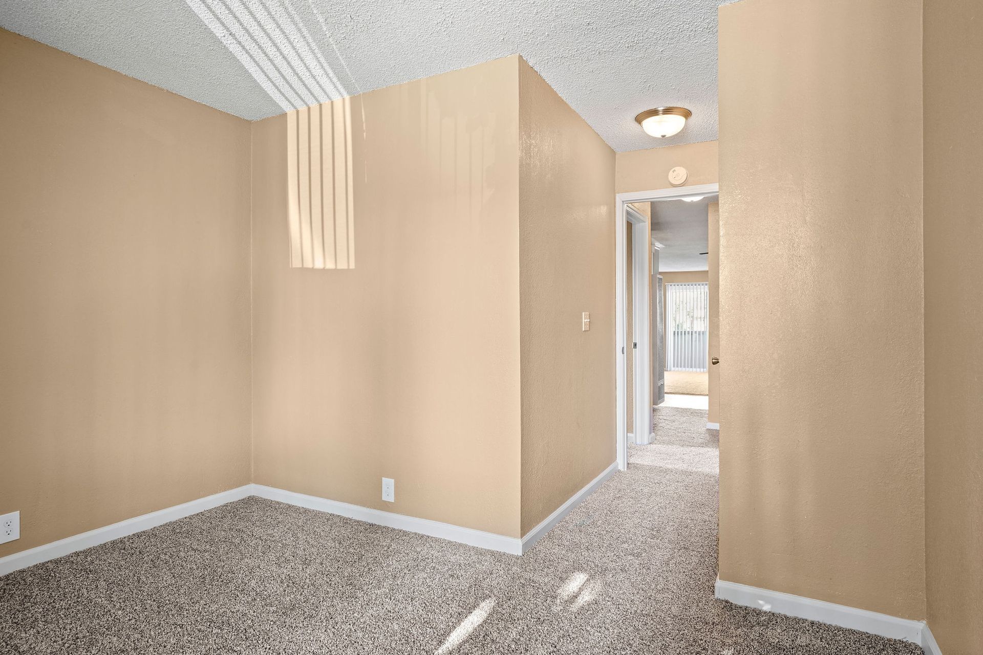 Empty beige hallway with carpeted floor and sunlight streaming through a distant doorway.