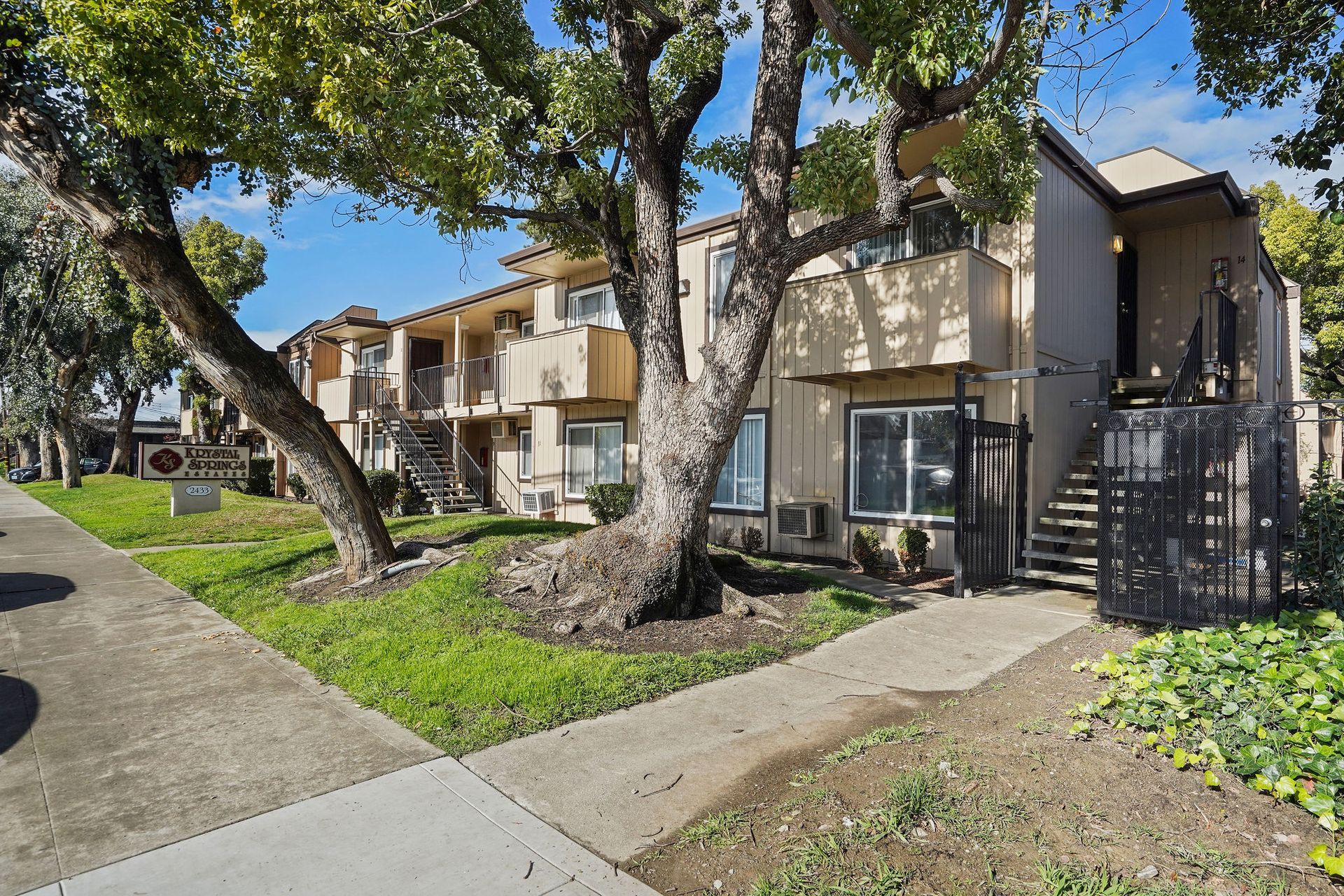 Apartment complex with balconies, large trees, and a sidewalk in front