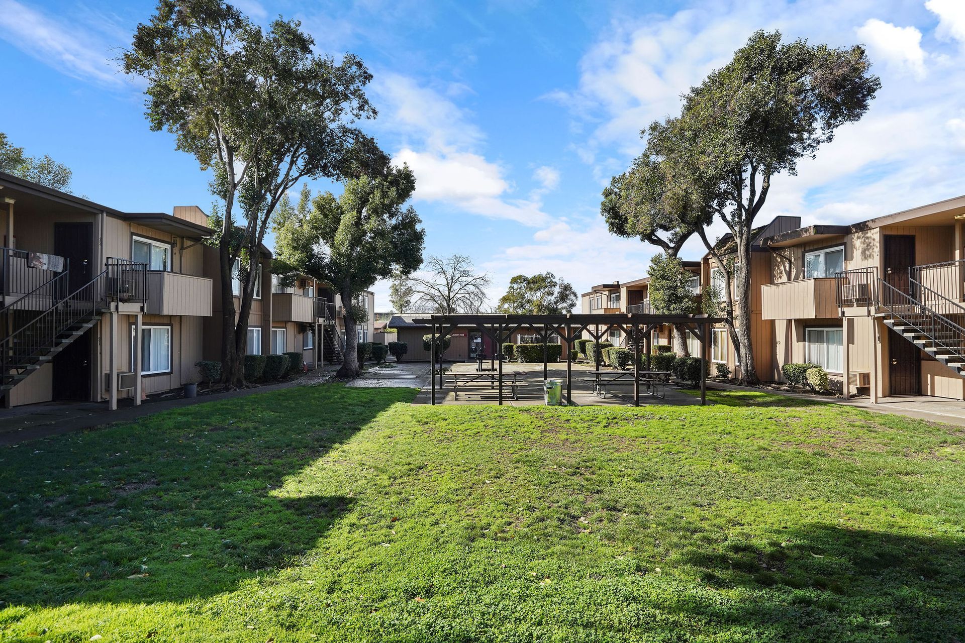 Apartment courtyard with green lawn, trees, and two-story buildings under a blue sky