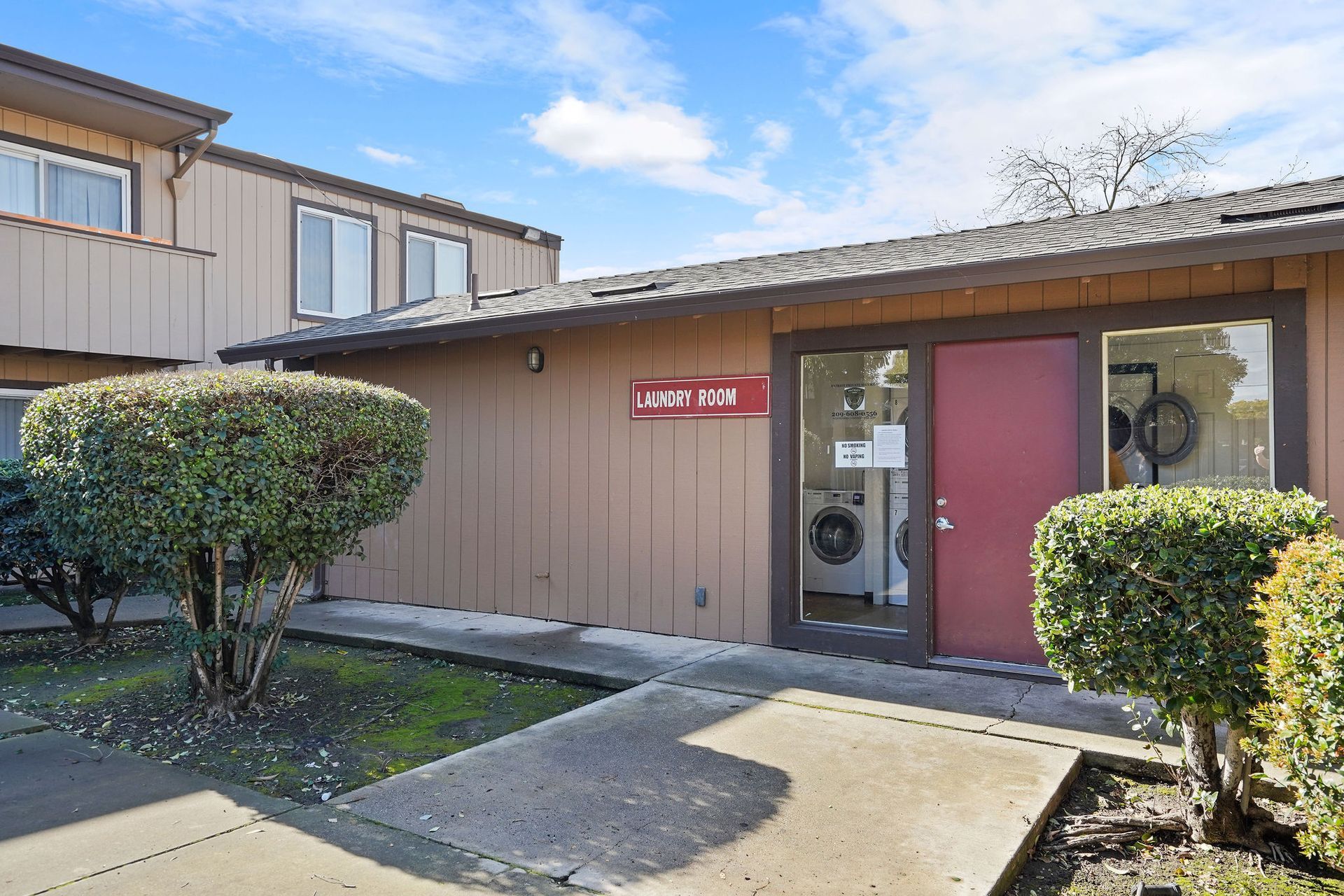 Apartment entrance with brown siding, red door, and trimmed bushes along a concrete walkway