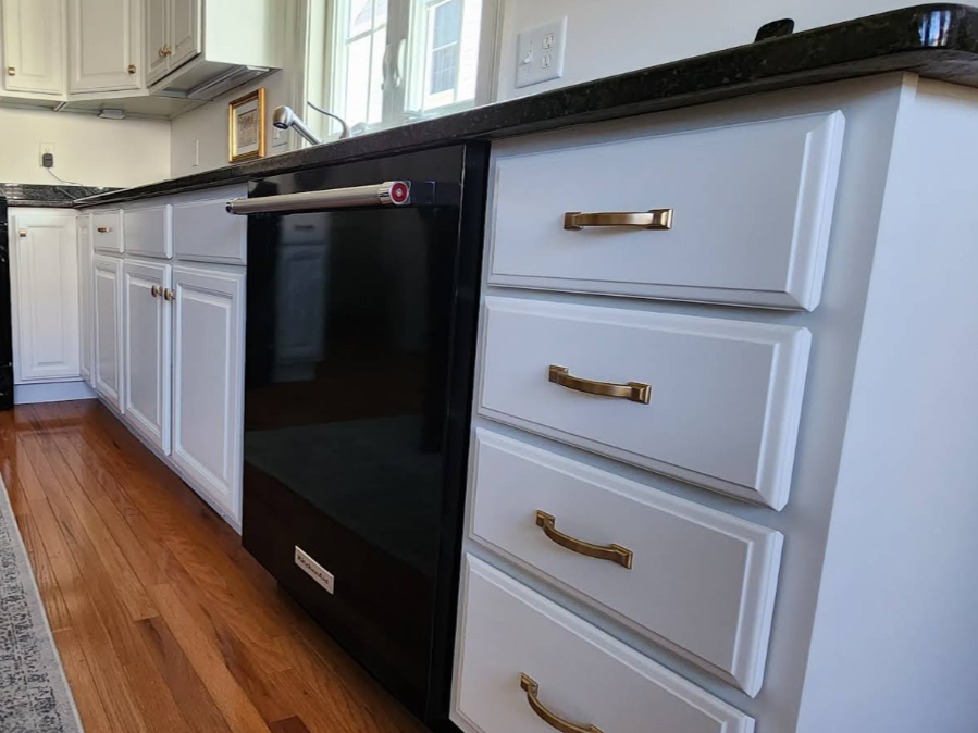 Black dishwasher between white kitchen cabinets and drawers with brass handles on a wood floor.