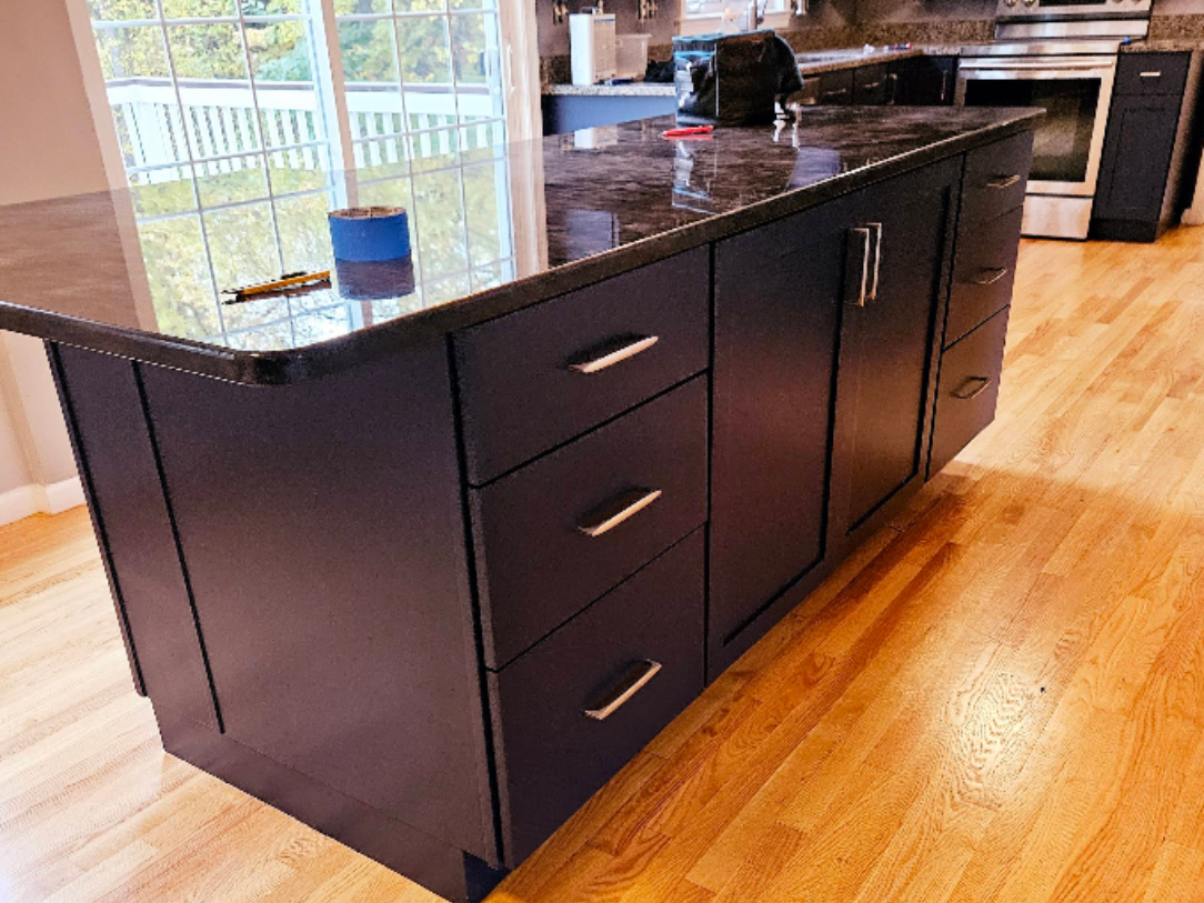 Dark granite kitchen island with black cabinets and drawers on a light wood floor.