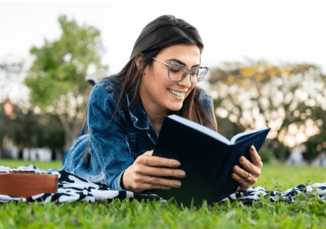 Una mujer está acostada sobre una manta en el césped leyendo un libro.