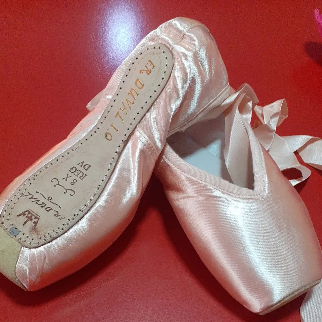 Zapatillas de punta de ballet de satén rosa pálido sobre una superficie roja, una de las zapatillas mostrando la suela.