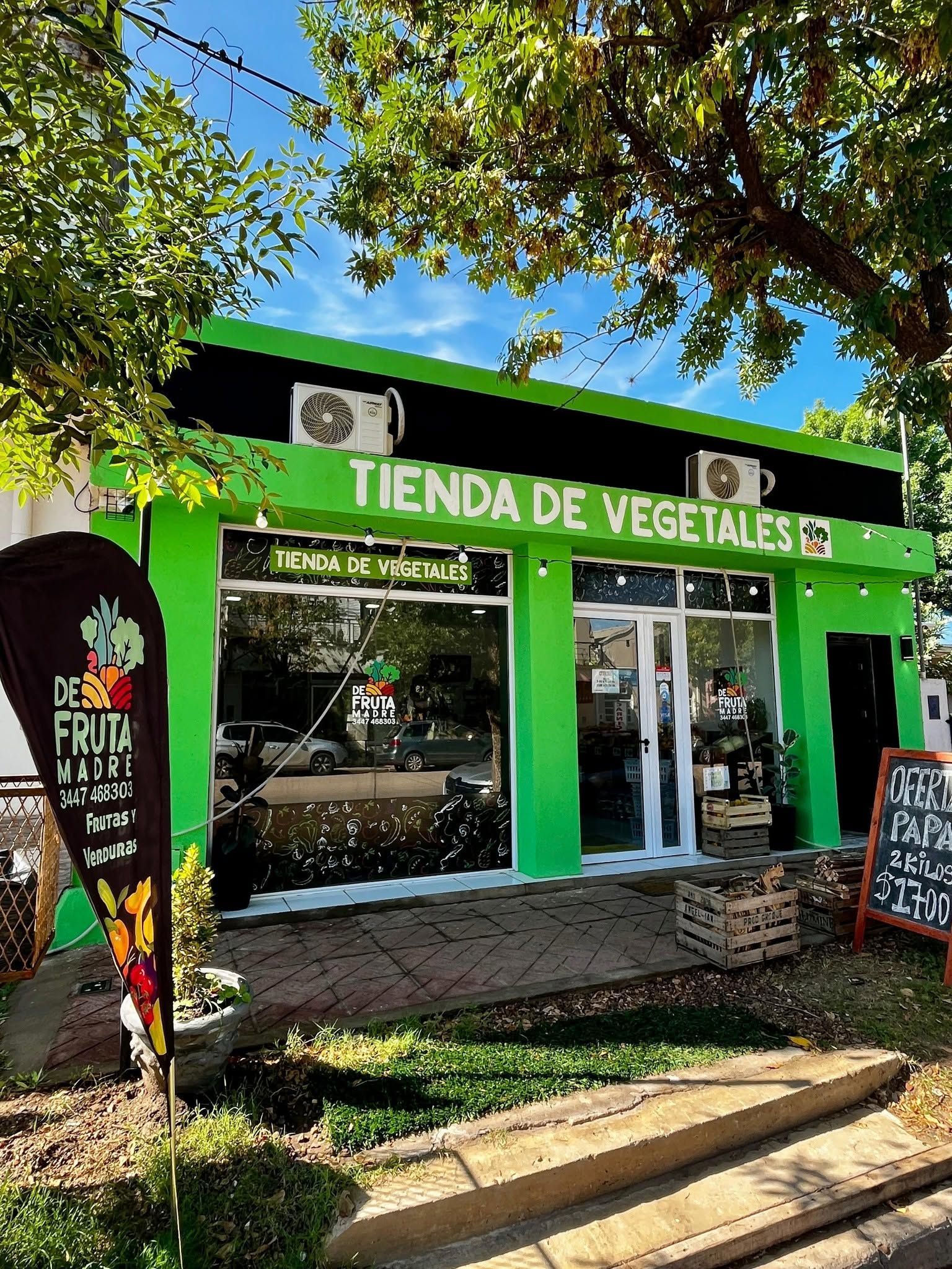 Fachada verde con el letrero “TIENDA DE VEGETALES” y carteles de productos, bajo árboles frondosos y un cielo azul.