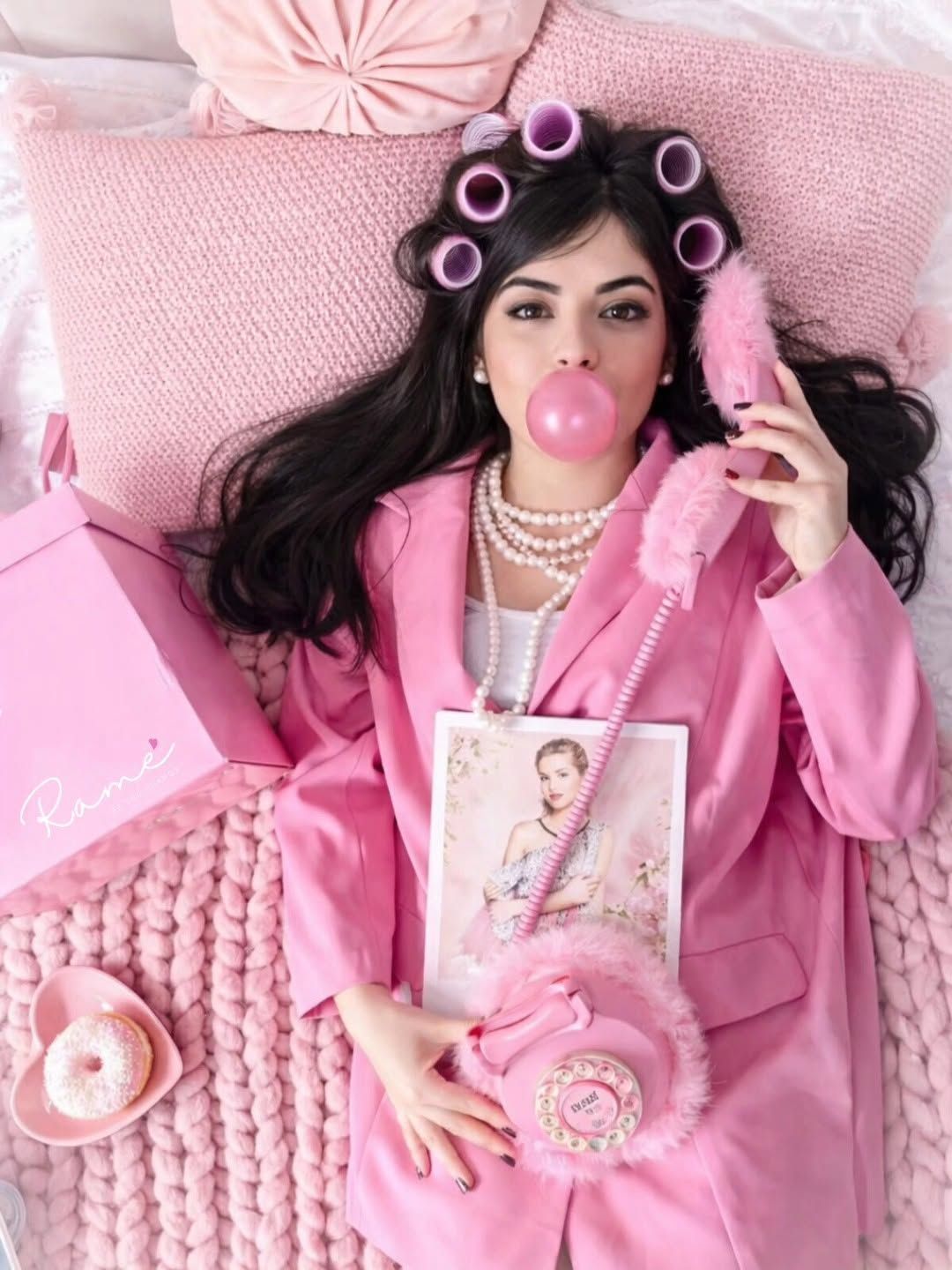 Mujer vestida de rosa, recostada en una cama, con rulos en el cabello y perlas, soplando una burbuja de chicle rosa.