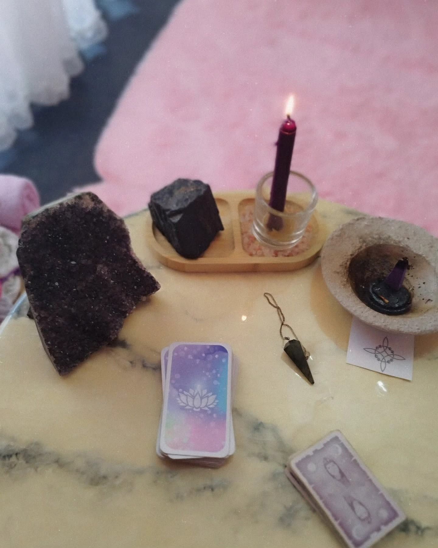 Pequeño altar con una vela encendida, cristales, cartas del tarot y un colgante sobre una superficie rosa esponjosa.