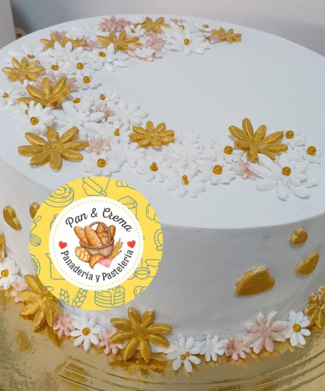 Pastel blanco con flores amarillas y rosa pálido y detalles dorados, decorado con un adorno con el logo de “Pan & Creme”.