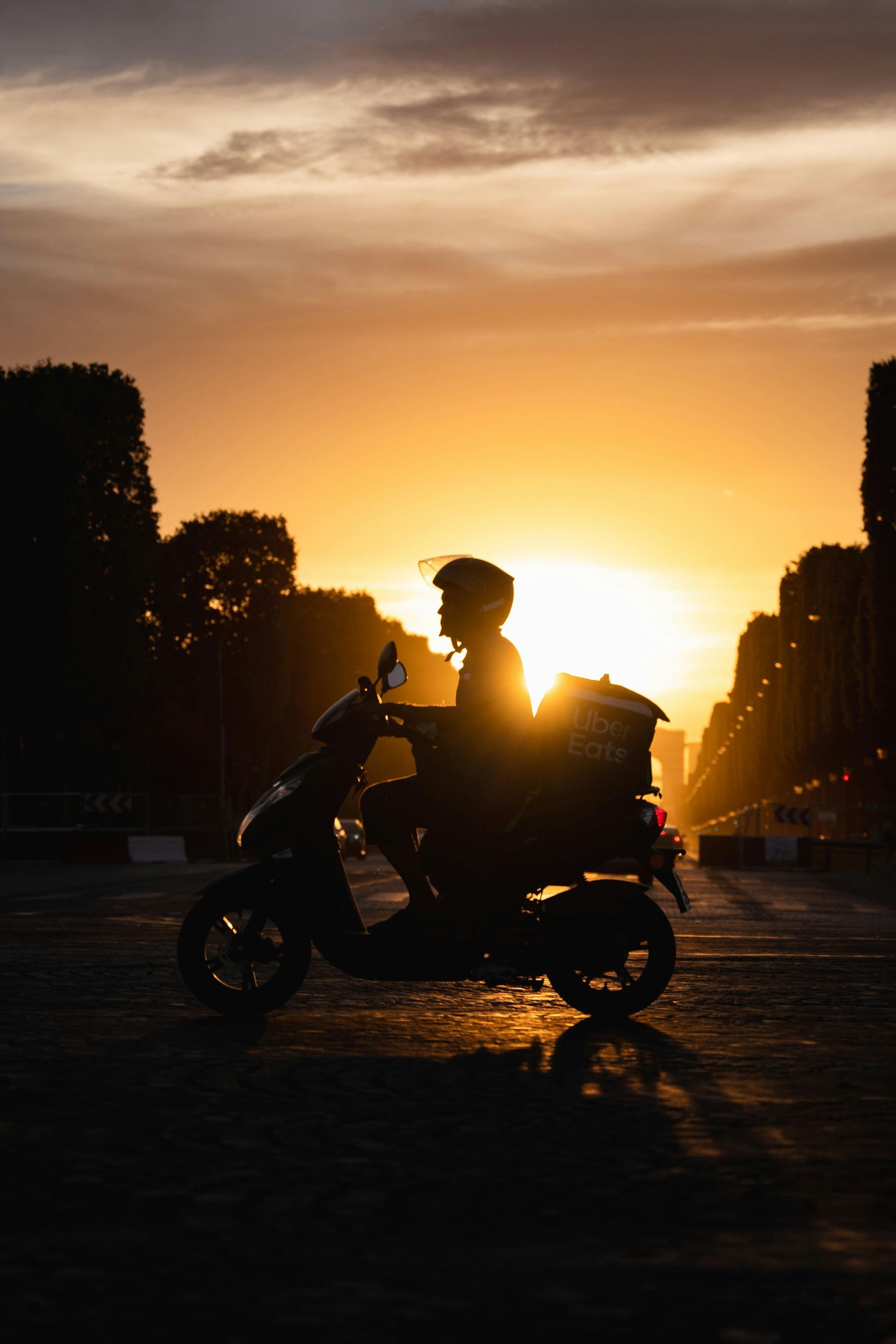 Silueta de dos personas en una moto al atardecer en una calle de la ciudad, con árboles que bordean la carretera.