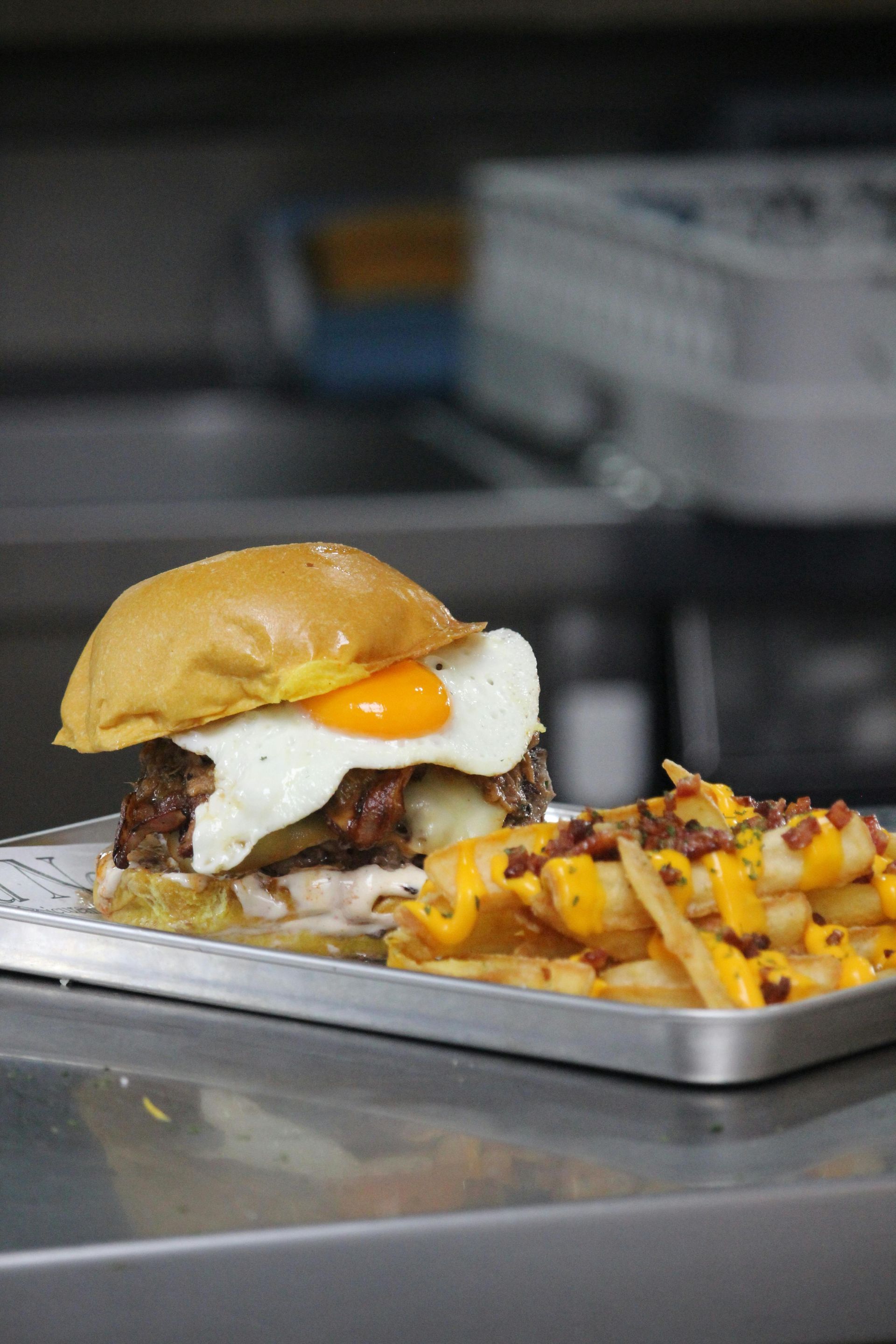 Hamburguesa con huevo frito y patatas fritas en una bandeja metálica en un entorno de cocina.