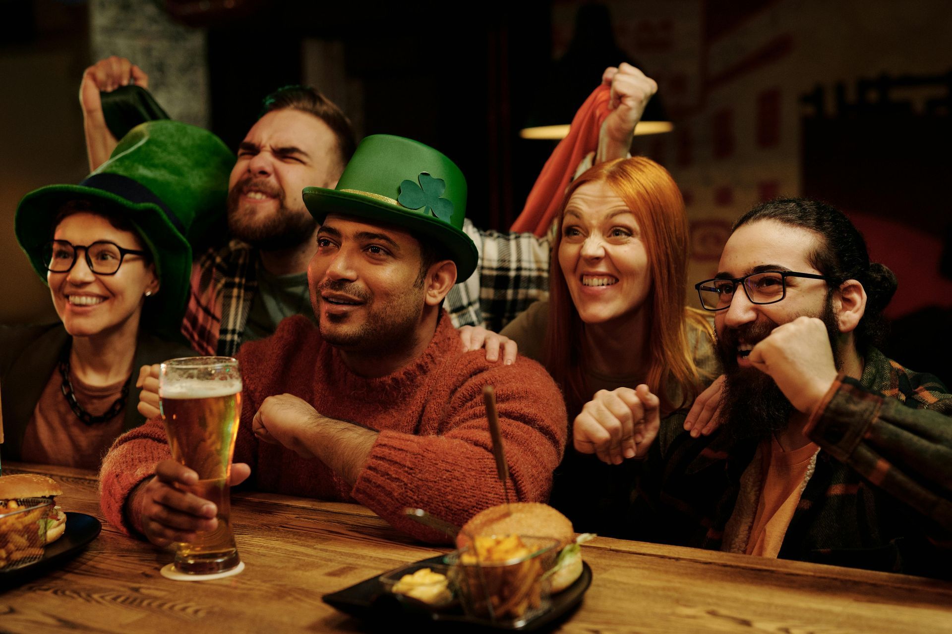 Amigos con gorros verdes y copas festivas brindan alrededor de una mesa de madera en un pub con iluminación cálida.