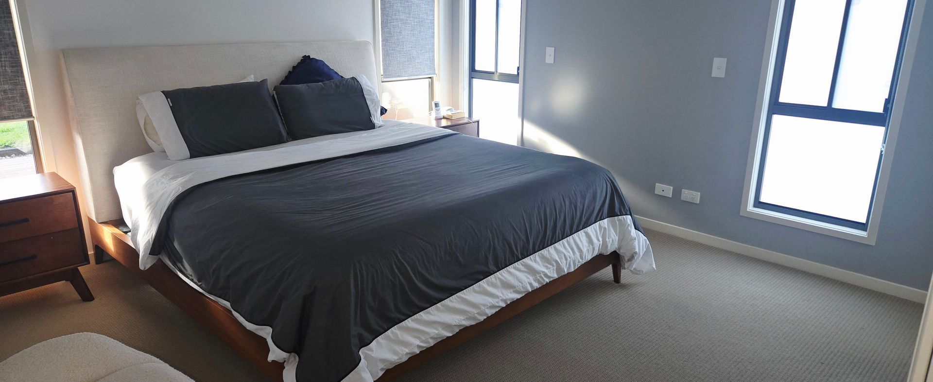 Neatly made bed with dark bedding in a bright bedroom with gray walls and two windows cleaned by elite care and clean.