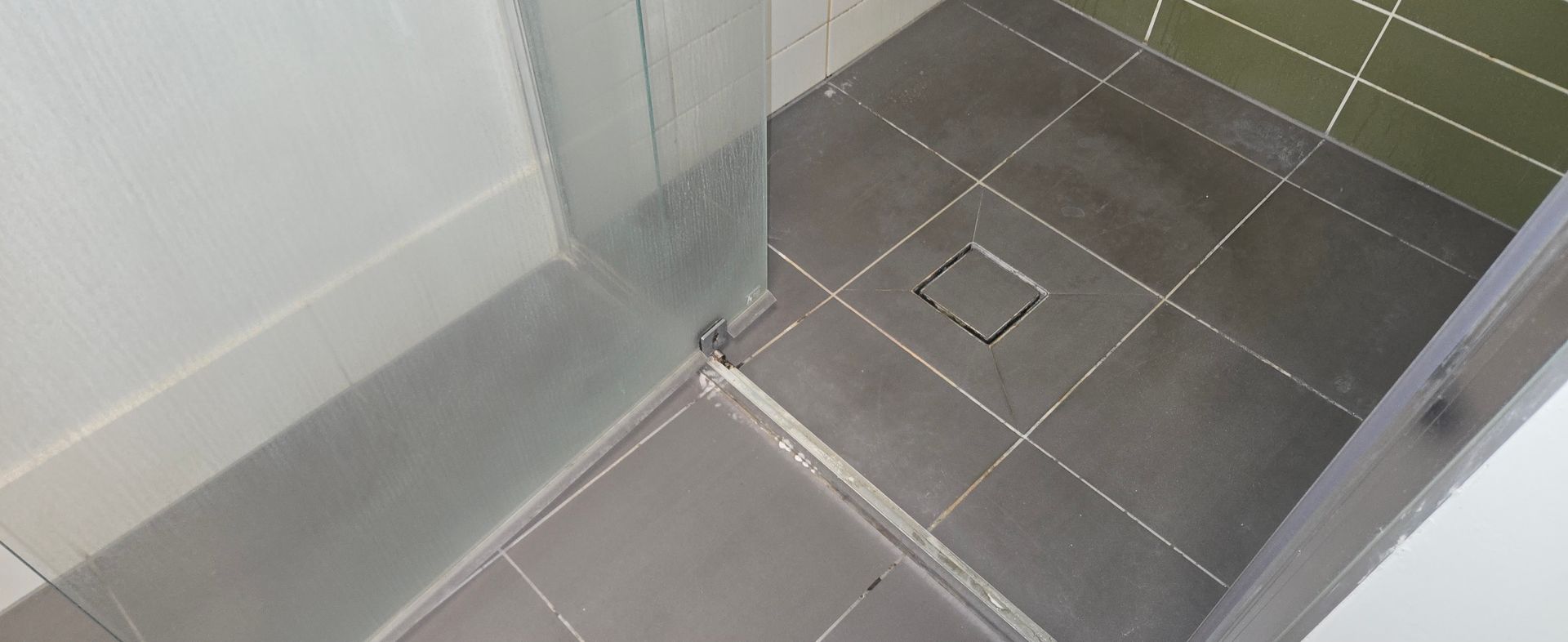 Dirty, calcium build up, tiled shower corner with glass partition and square floor drain. 