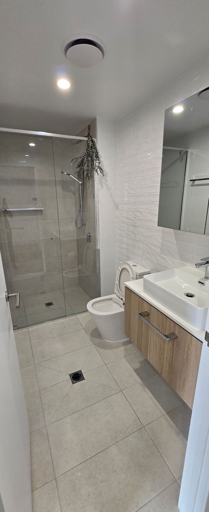 Modern bathroom with glass shower, white tile walls, wood vanity, sink, and toilet. Furnished clean apartment in Surfers Paradise