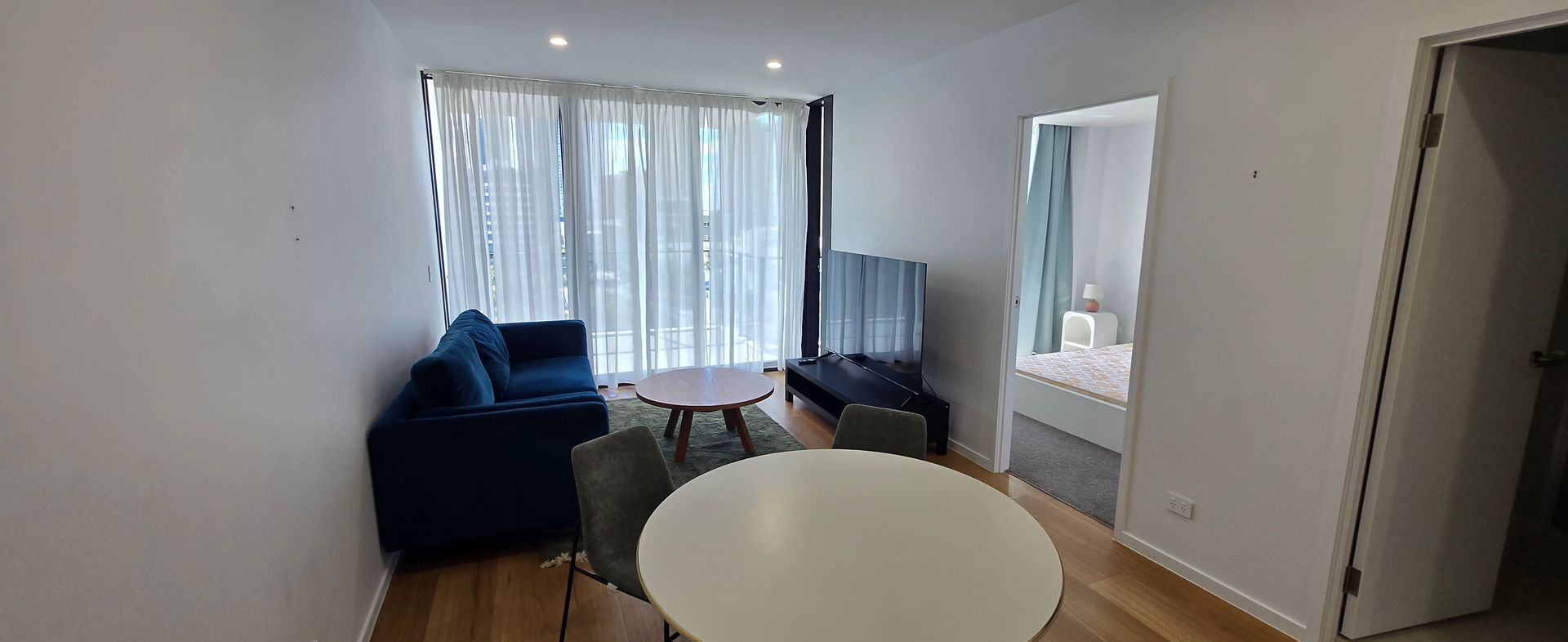 Bright living room with blue sofa, round table, window blinds, and open doorway. Furnished clean apartment in Surfers Paradise