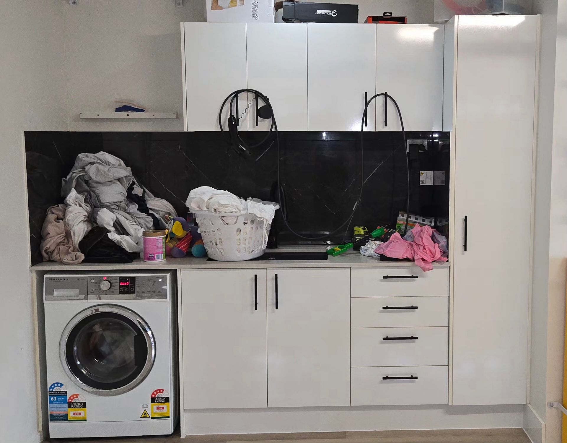 Cluttered kitchen with white cabinets, black backsplash, washing machine, and sink under hanging items.