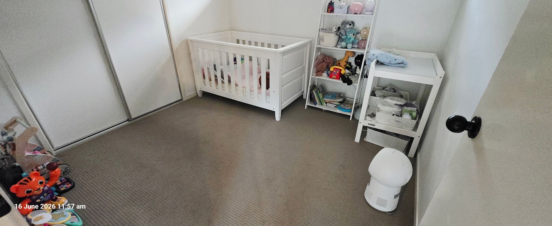 Empty nursery with a white crib, toy shelves, and a small white stool on beige carpet.