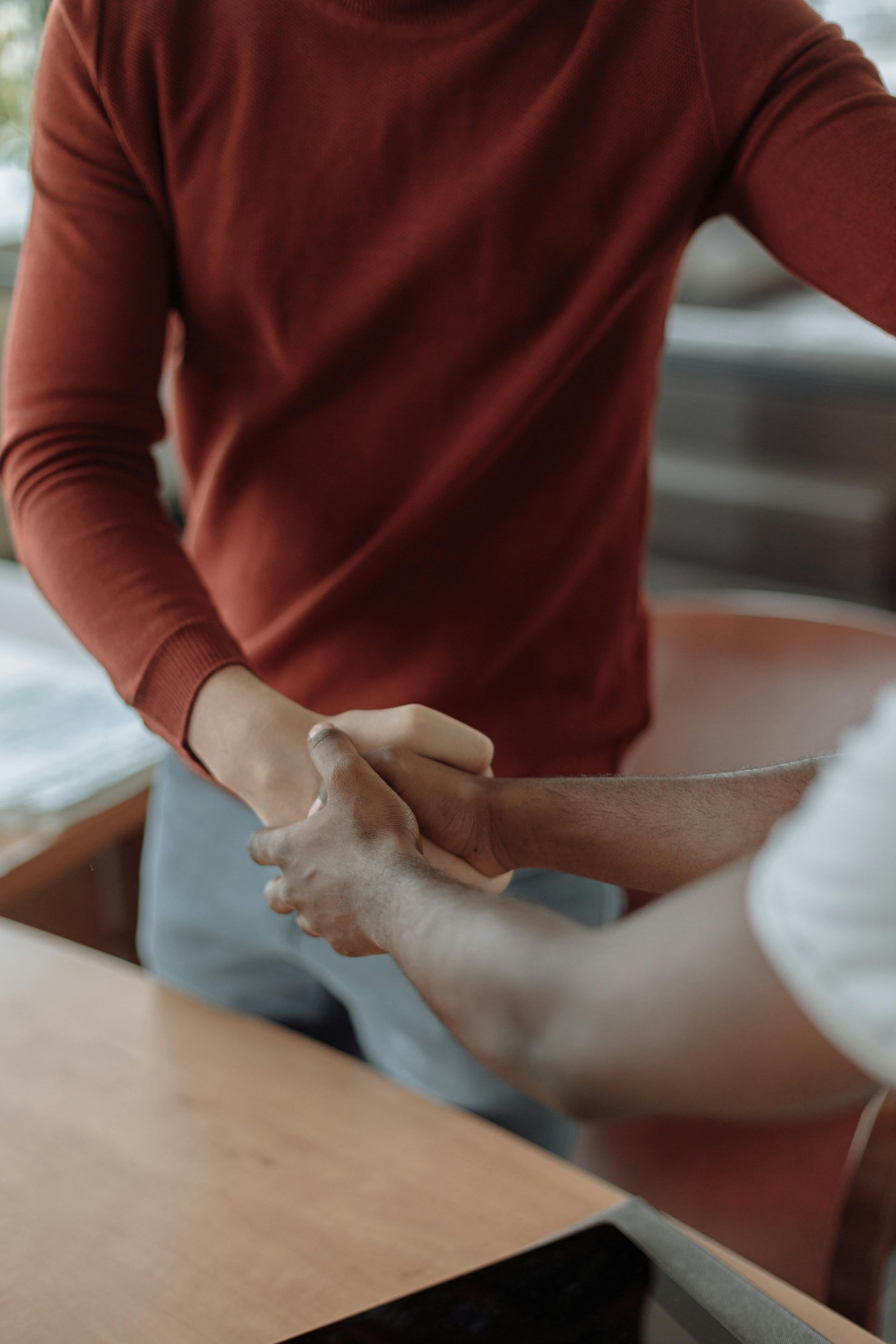 Two people shaking hands across a table, one in a rust sweater in an office setting