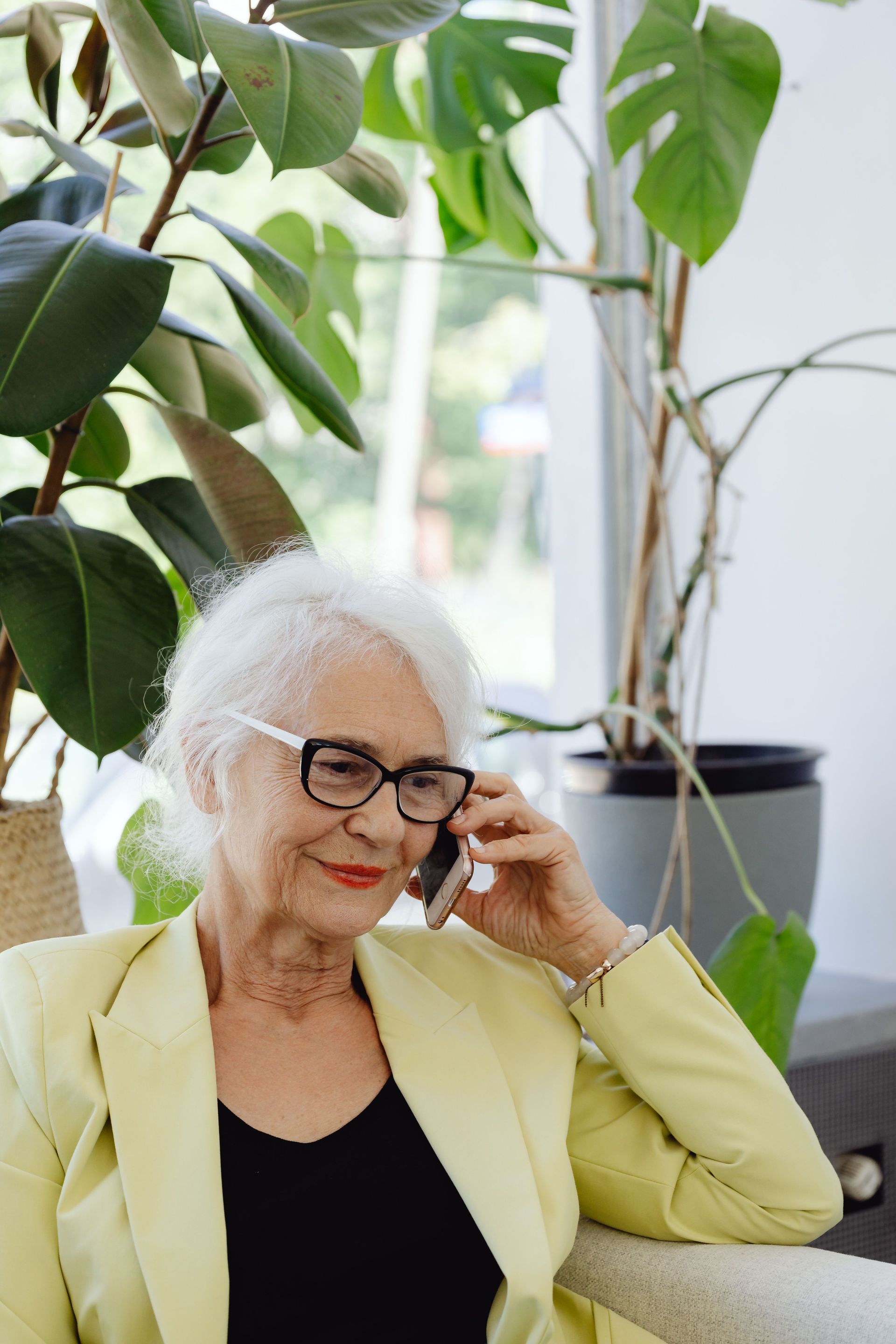 Woman with white hair and glasses sitting indoors by a window, holding a phone and smiling in a light yellow blazer