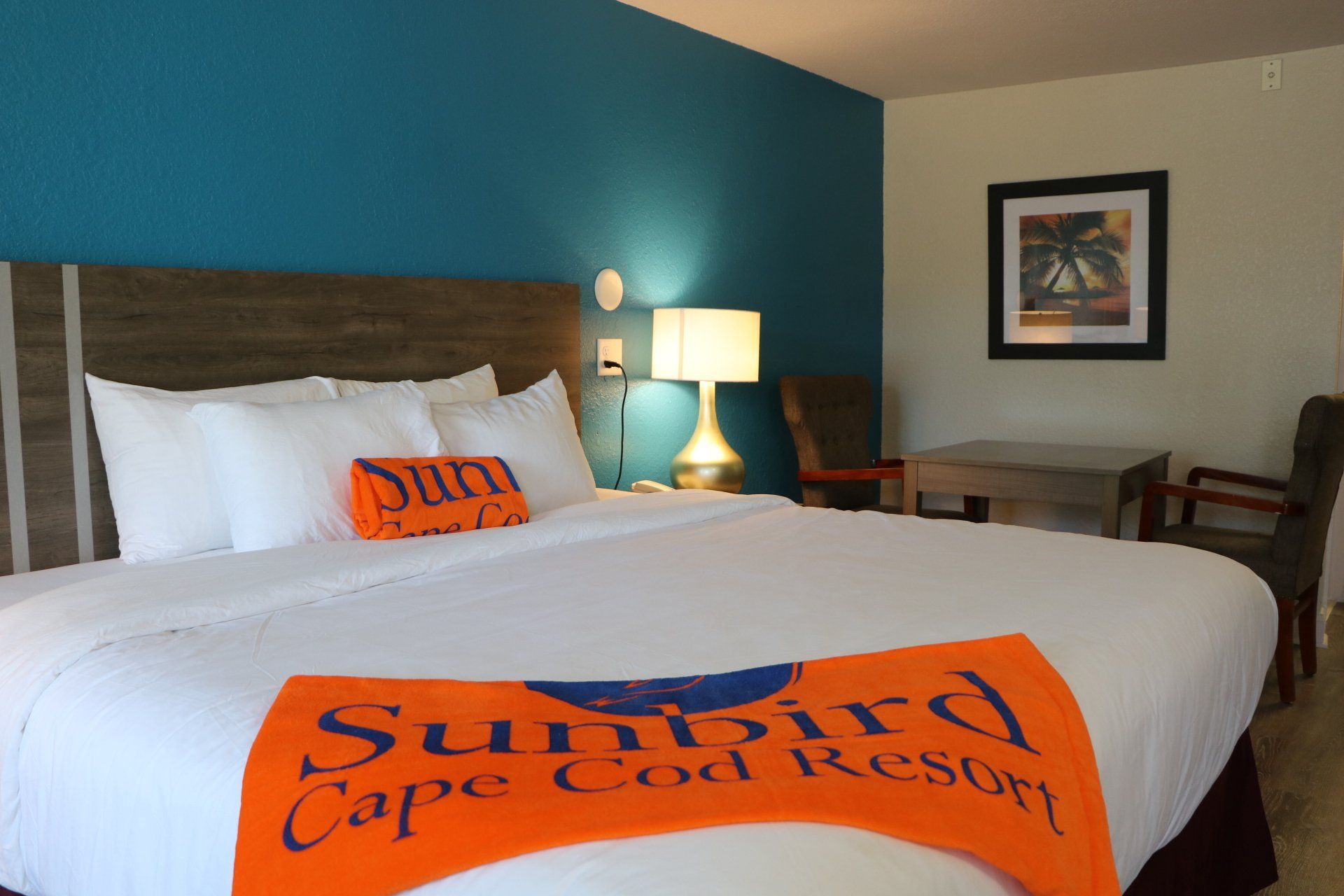 Hotel room with king bed, teal wall, orange Sunbird resort towels, lamp, artwork.