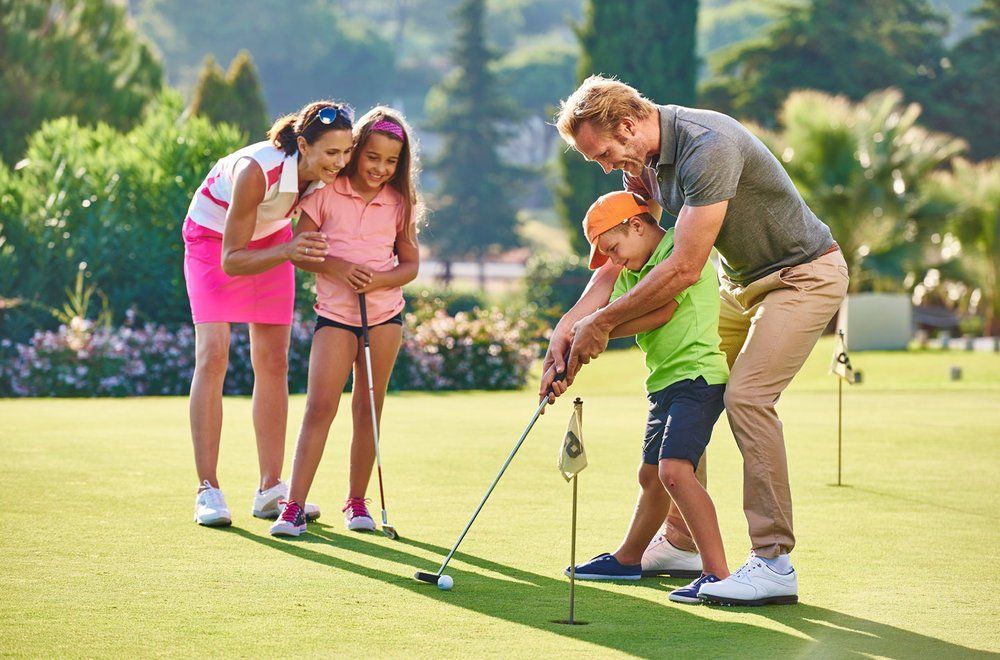 Family on a golf course: Dad teaches son to putt, mom & daughter watch, sunny day.