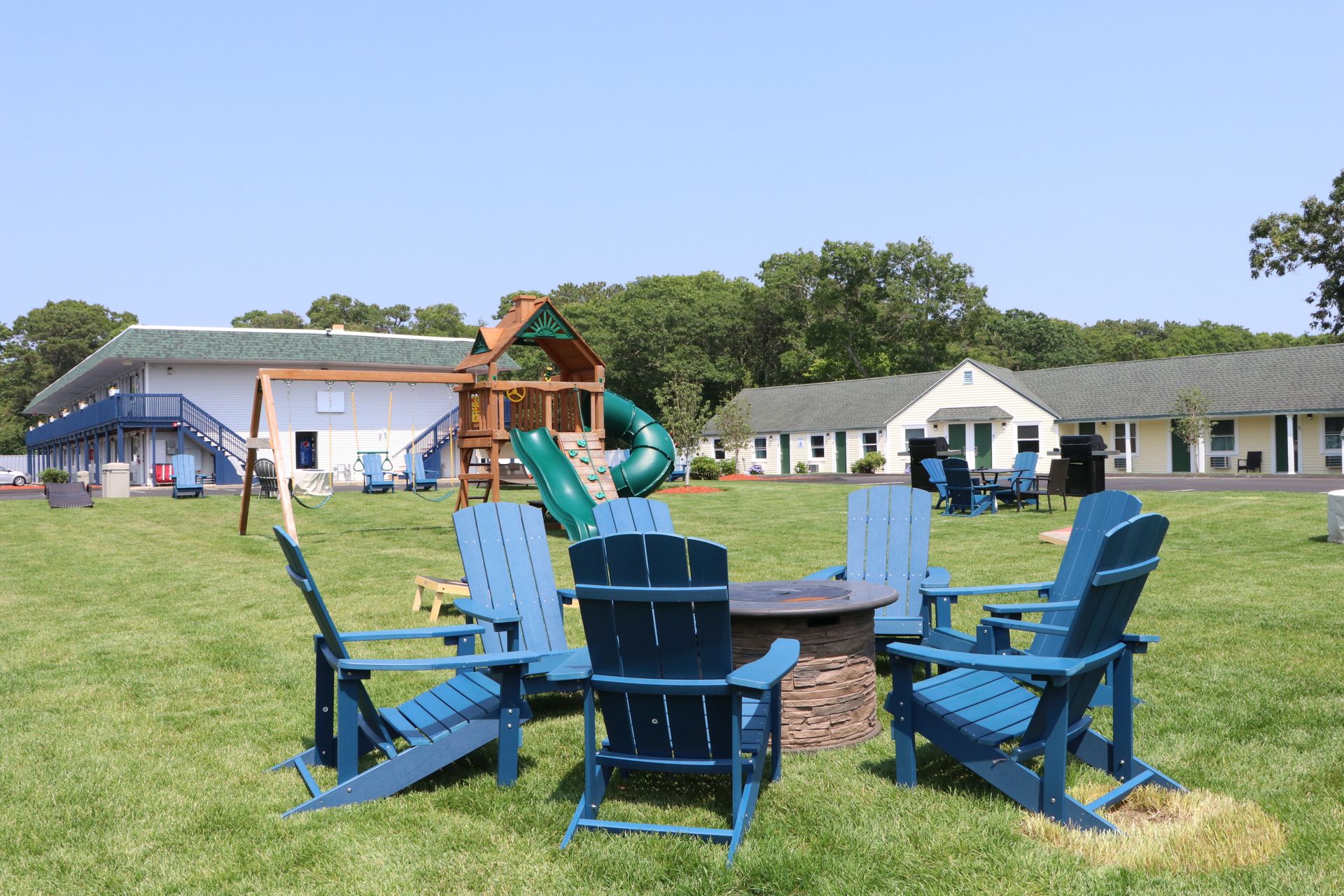 Blue Adirondack chairs around a fire pit on a grassy lawn; a playground and motel buildings in the background.
