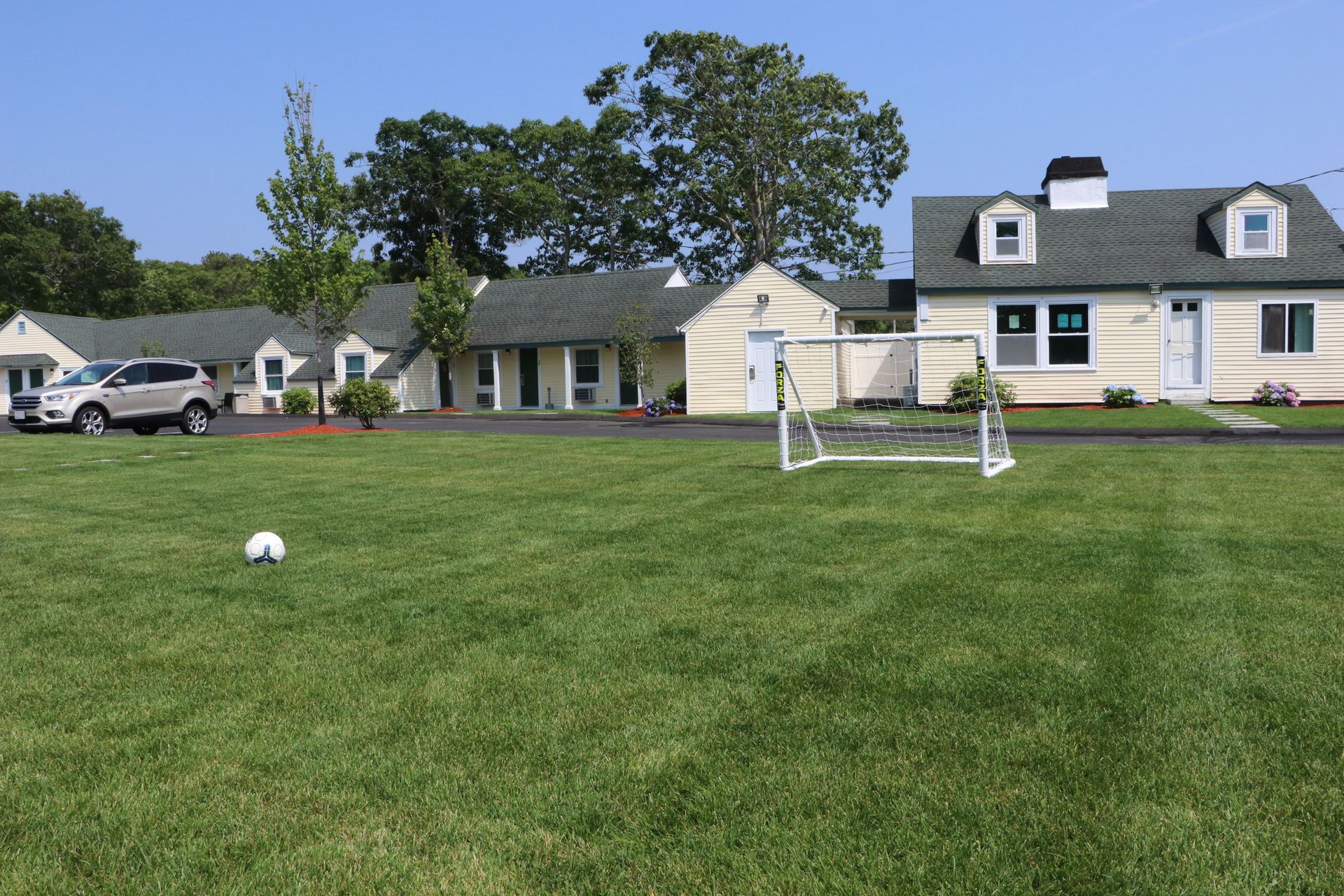 Soccer ball on grass field in front of a light yellow building with green roof, soccer goal, and car.