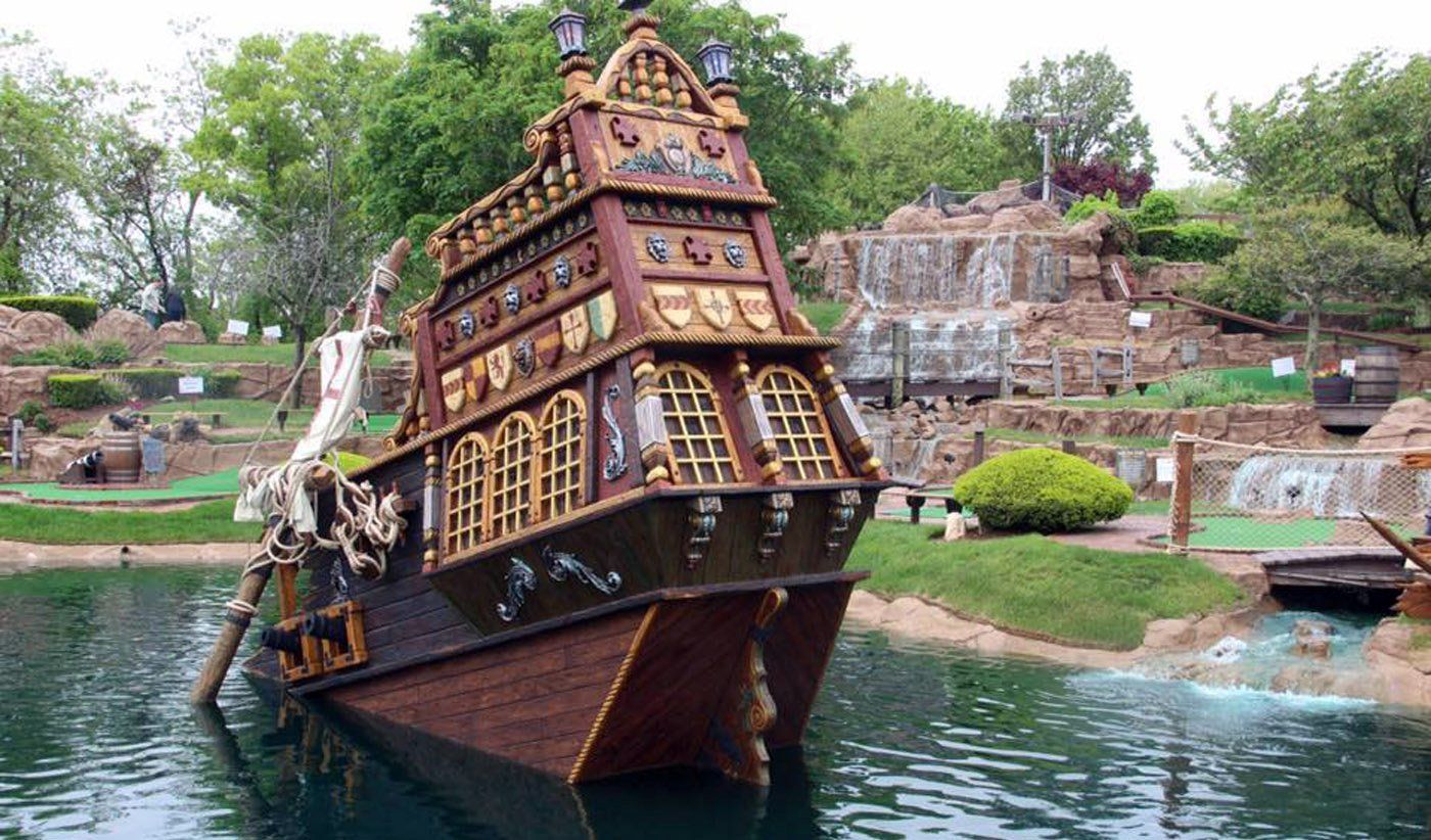 Pirate ship-themed miniature golf course. Brown wooden ship, waterfall, green grass, and trees in background.