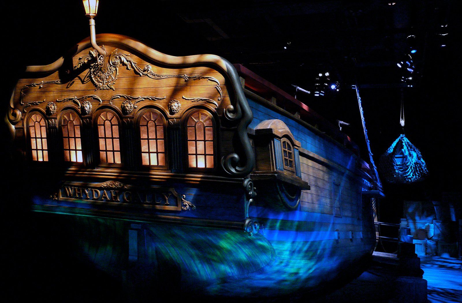 Stern of a blue and gold ship with decorative windows, illuminated against a dark background, projected water effect.