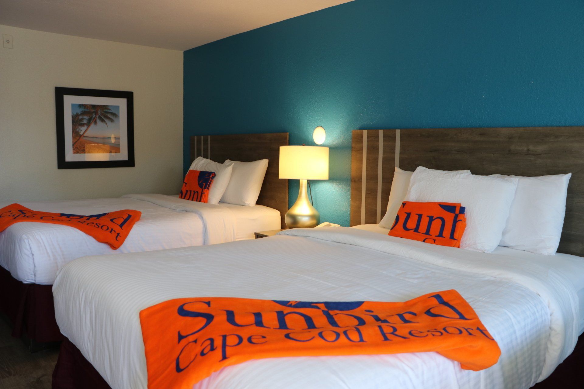 Two queen beds in a hotel room with orange Sunbird Cape Cod Resort blankets and turquoise accent wall.