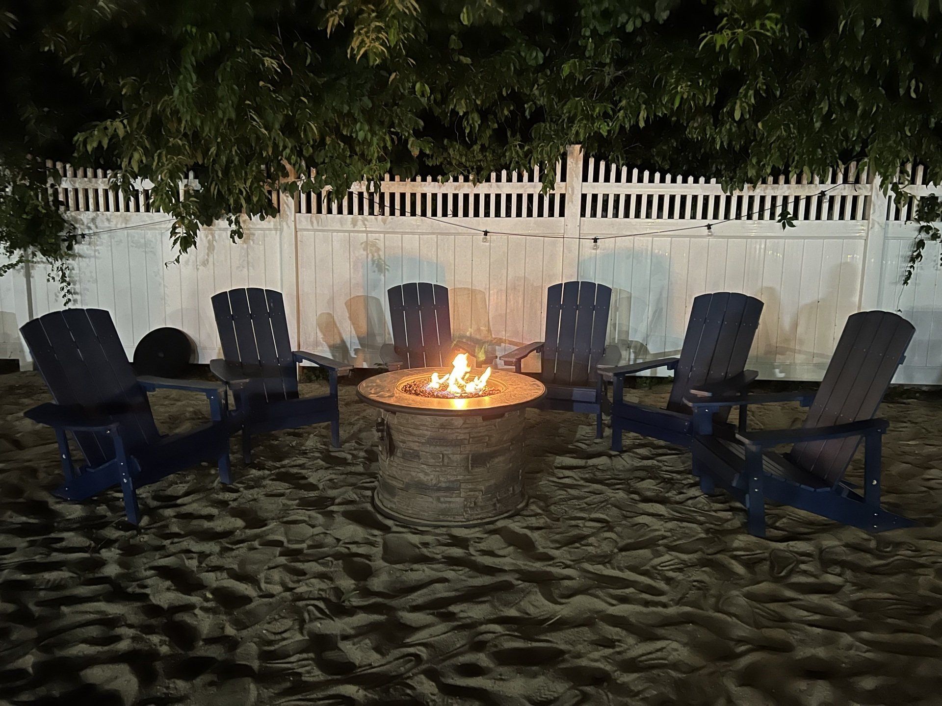 Six blue chairs circle a fire pit on sand, in front of a white picket fence at night.
