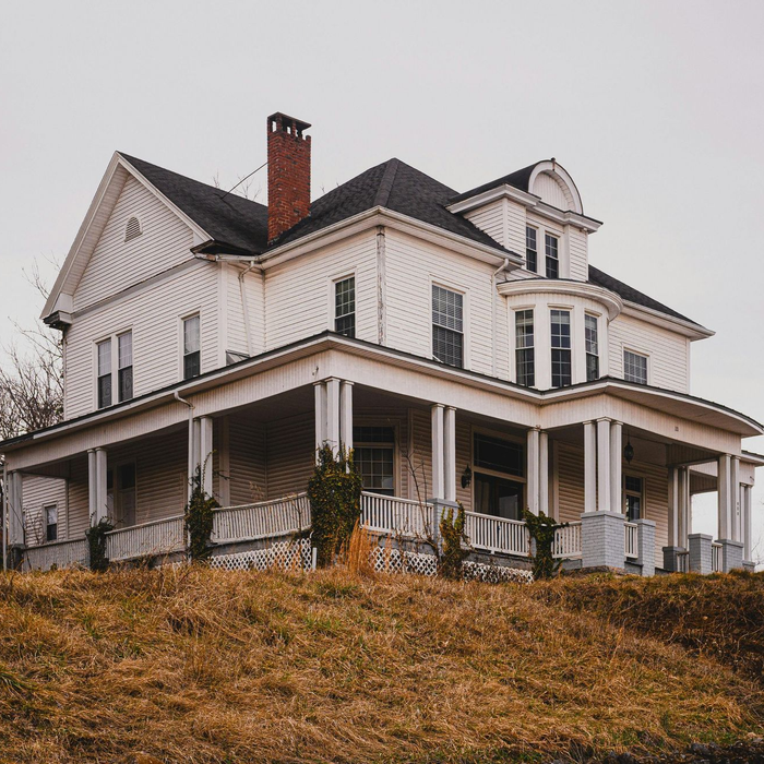 Large white Victorian house with a wraparound porch on a grassy hill under an overcast sky in idaho falls